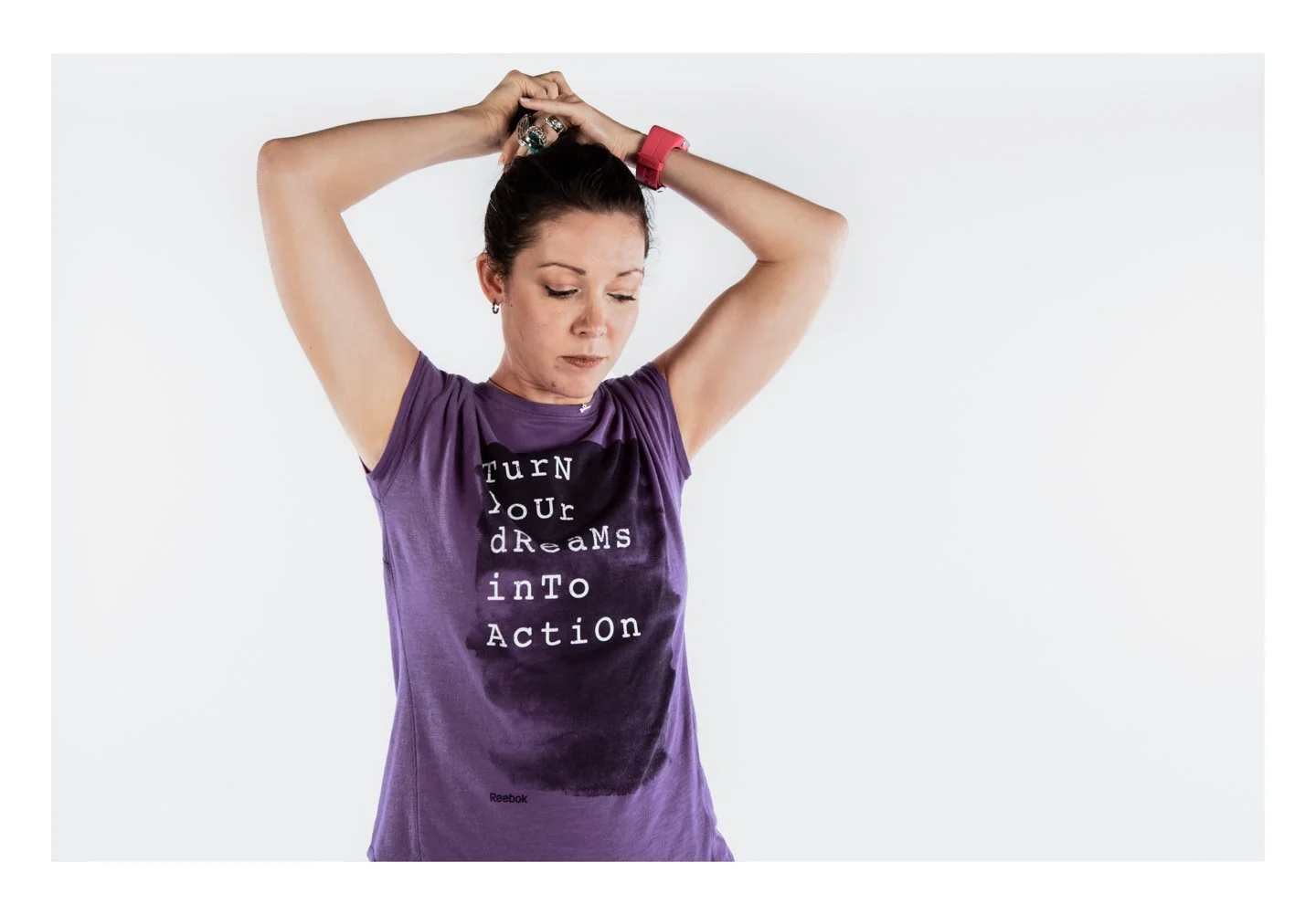 A woman in a purple t-shirt with the words "Turn Your Dreams into Action" is stretching her arms above her head. She has her eyes closed and short dark hair.
