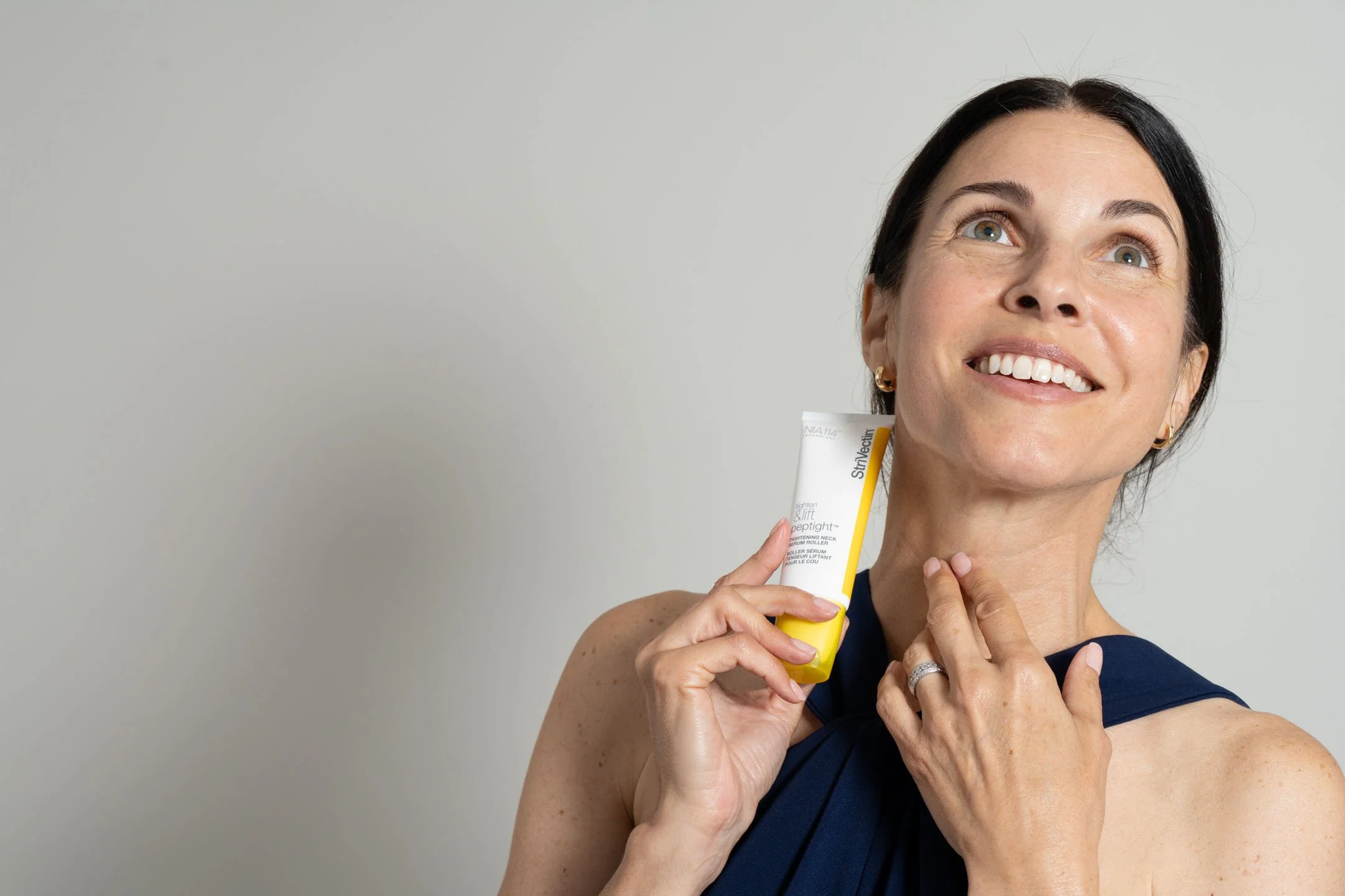 A woman with dark hair, wearing a navy sleeveless top, smiling and holding a yellow and white tube of skincare cream near her face, against a plain light background.