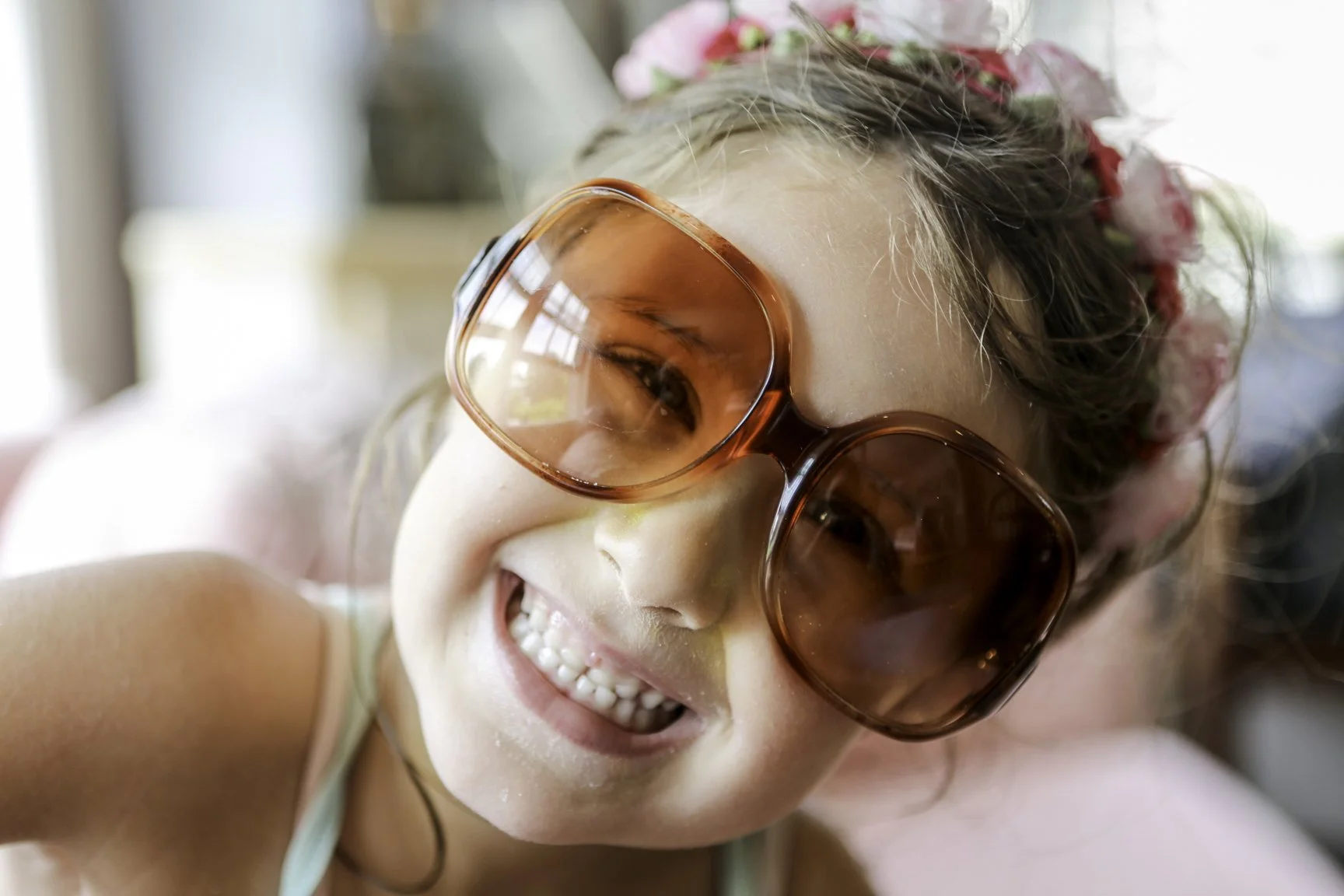 A young girl wearing large, orange-tinted sunglasses and a flower headband, smiling brightly.