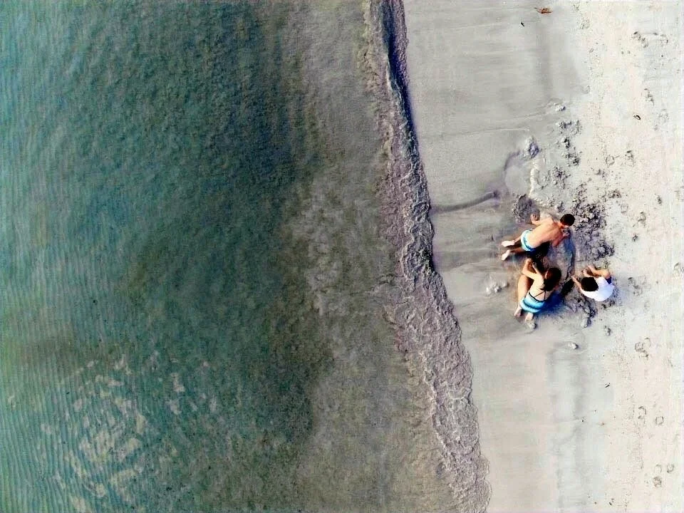 Four children playing on the sandy beach near the water, with one child digging in the sand and others standing nearby.