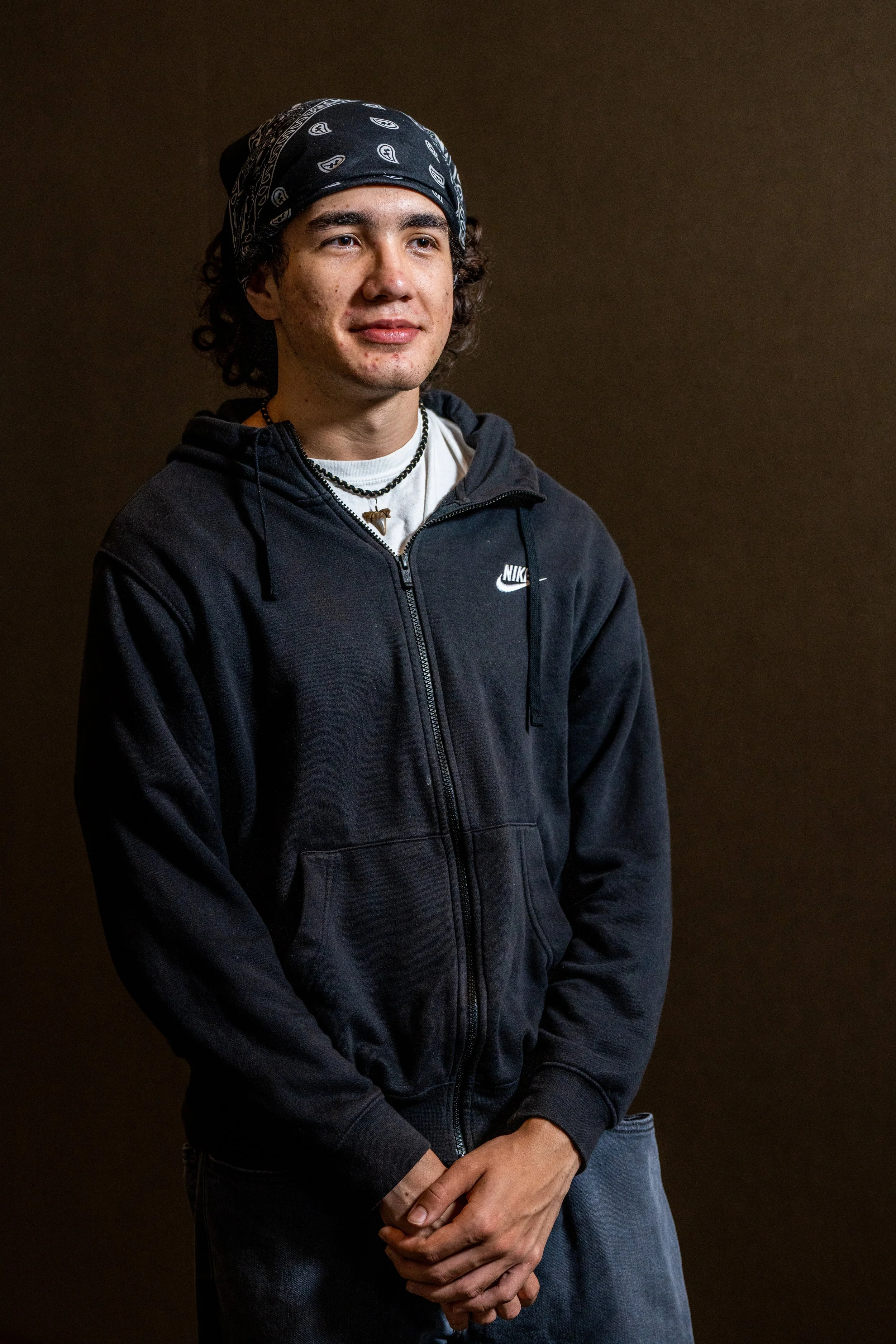 A young man with curly hair, wearing a black bandana, a black Nike hoodie, and blue jeans, standing in front of a plain brown background.
