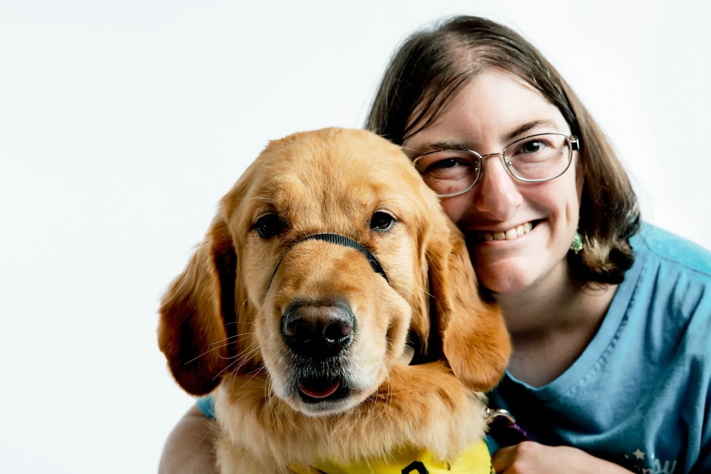 A smiling woman with glasses hugging a golden retriever puppy with a harness, both facing the camera against a white background.