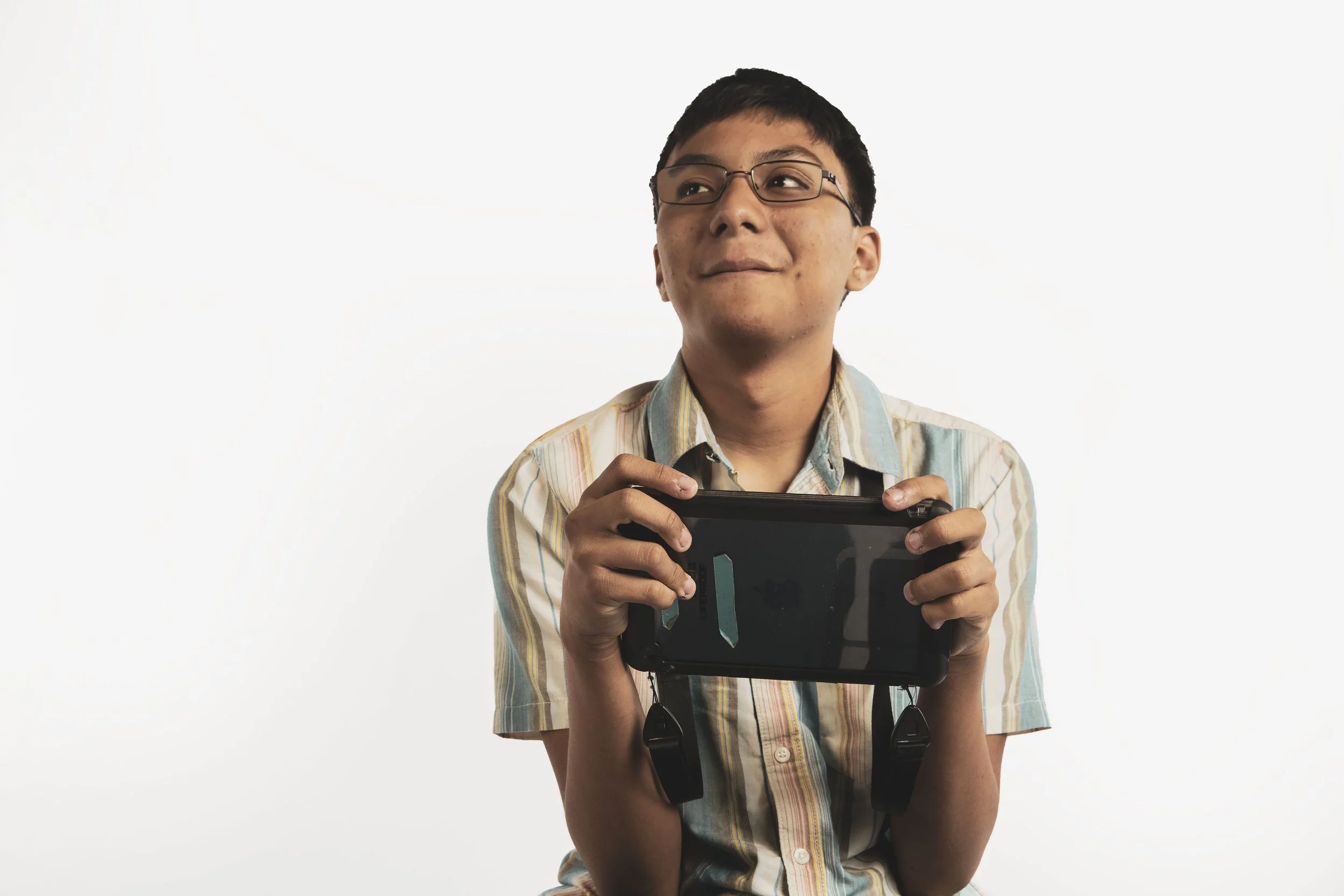 A young man with glasses holding a handheld gaming device, dressed in a short-sleeved striped shirt, looking off to the side with a thoughtful expression, against a plain white background.