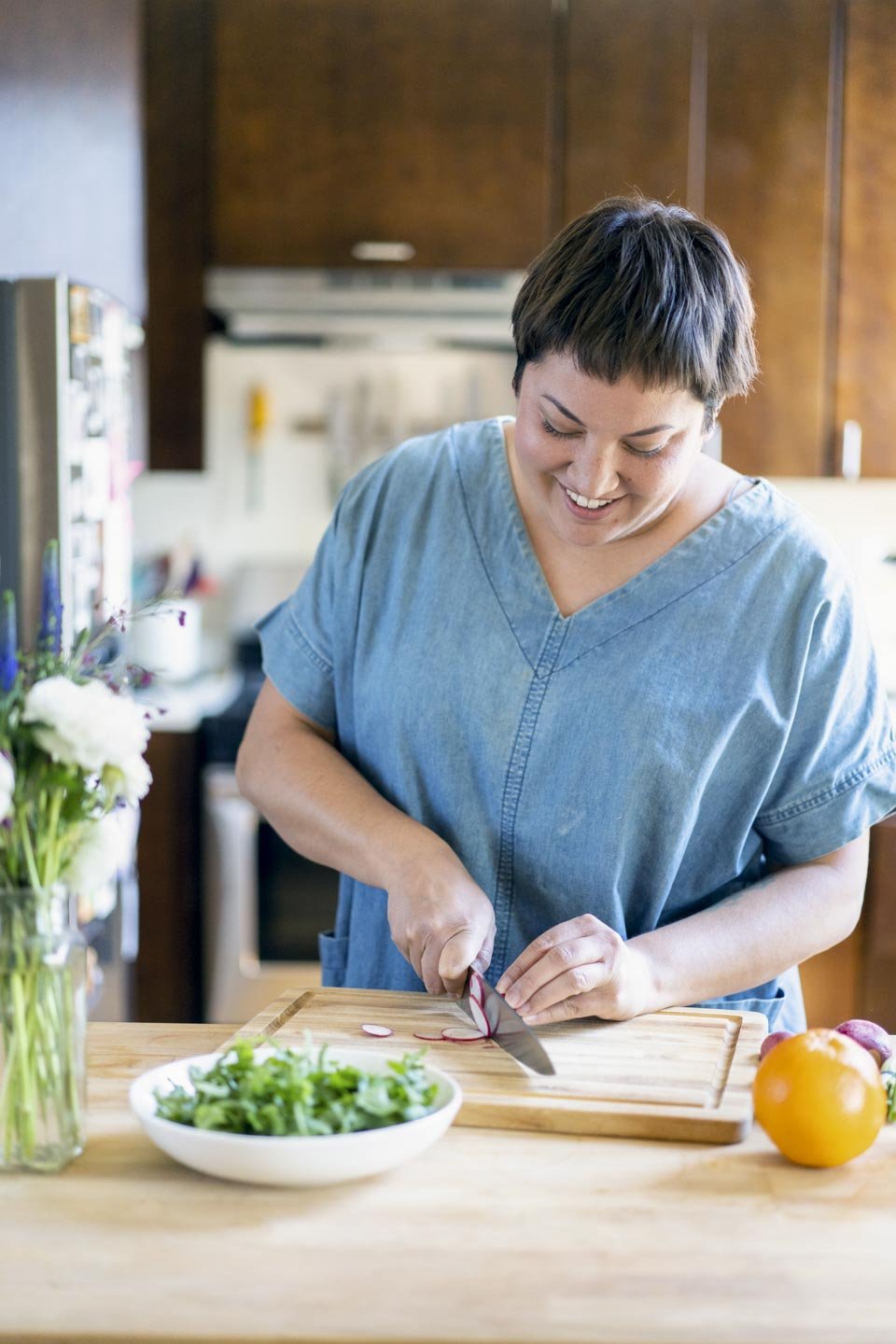 A person with short brown hair, wearing a blue scrub top, is smiling while slicing a radish on a wooden cutting board. A bowl of green salad and other vegetables are visible on the kitchen counter.