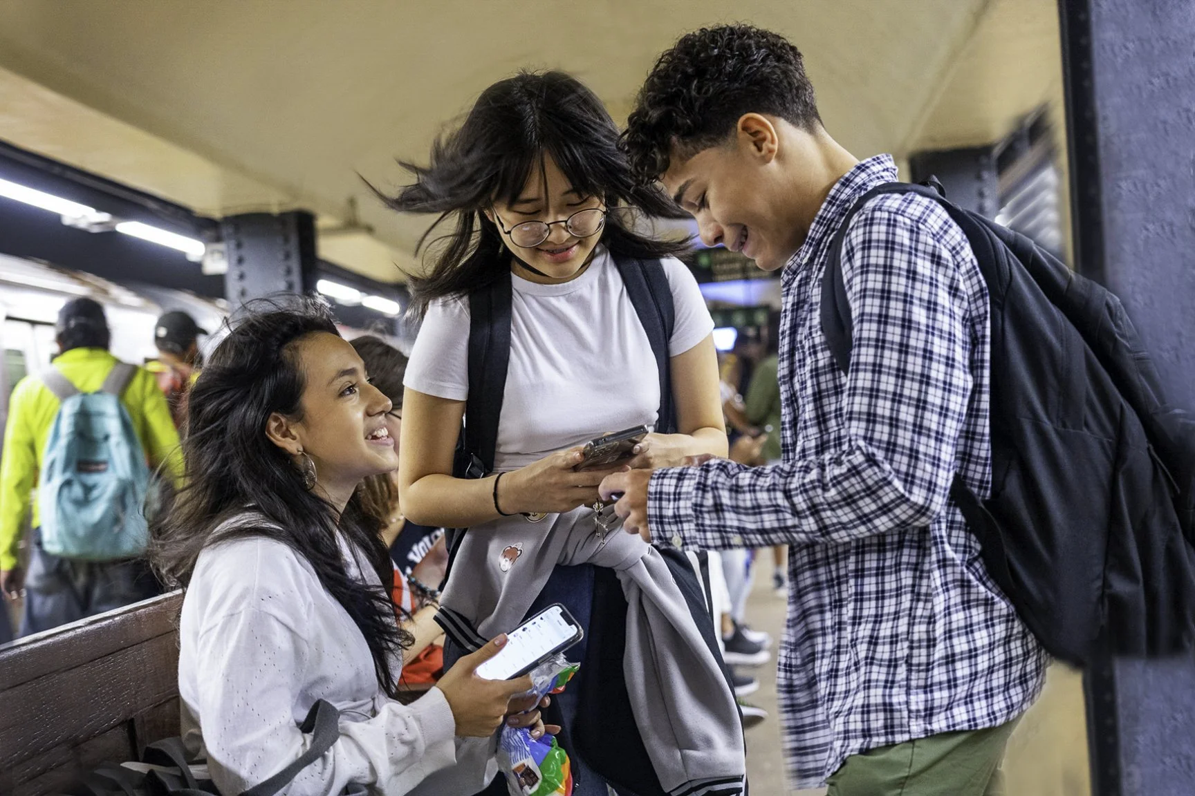 Three teenagers, two girls and one boy, at a subway station looking at a smartphone and smiling. The girl sitting on the bench holds a phone, the girl standing shows a phone to the boy, all wearing backpacks.