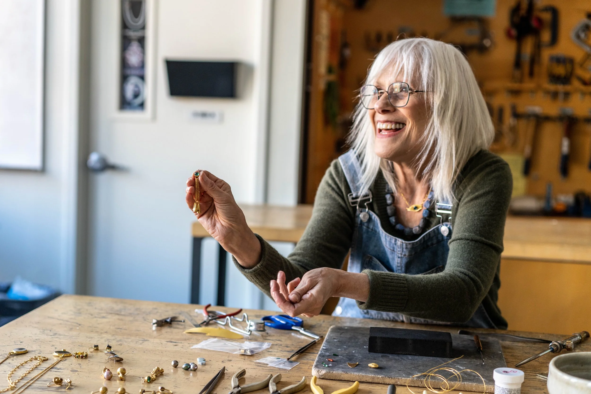 A cheerful elderly woman with gray hair, glasses, and a green sweater working on jewelry in a workshop. She is holding a small piece of jewelry, smiling and looking to the side, surrounded by tools and jewelry supplies on a wooden table.