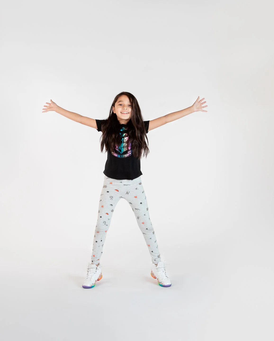 A girl with long dark hair standing with her arms outstretched, smiling, wearing a black t-shirt and light gray pants with small colorful patterns, and white sneakers with rainbow-colored soles.