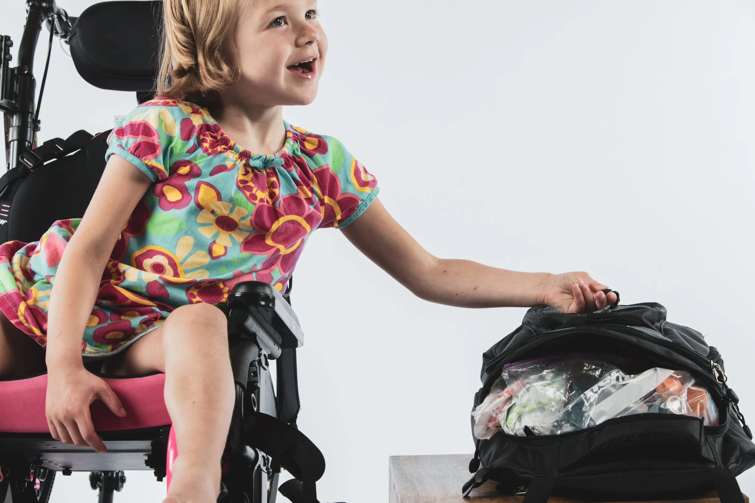 Young girl with a smile in a colorful floral dress reaching into a black backpack on a table, sitting in a pink and black wheelchair.