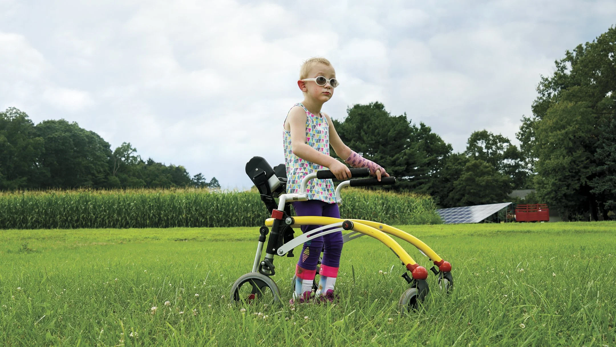 A young girl with a cast on her arm standing on a grass field next to a yellow and black walker with wheels, wearing sunglasses, colorful socks, and a geometric patterned dress, with a farm or rural area in the background.