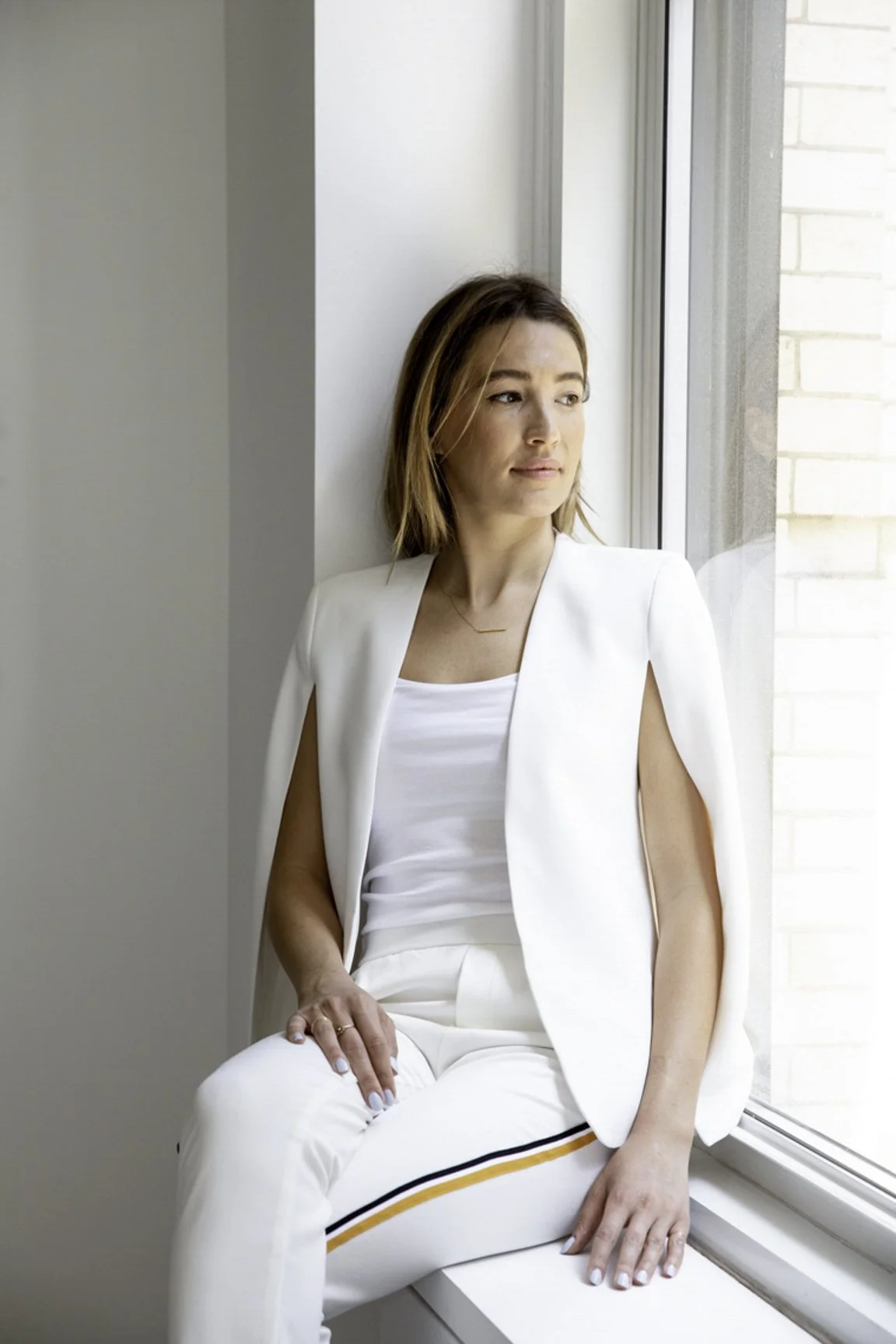 Woman sitting on windowsill looking out the window, wearing a white sleeveless outfit with a blazer.