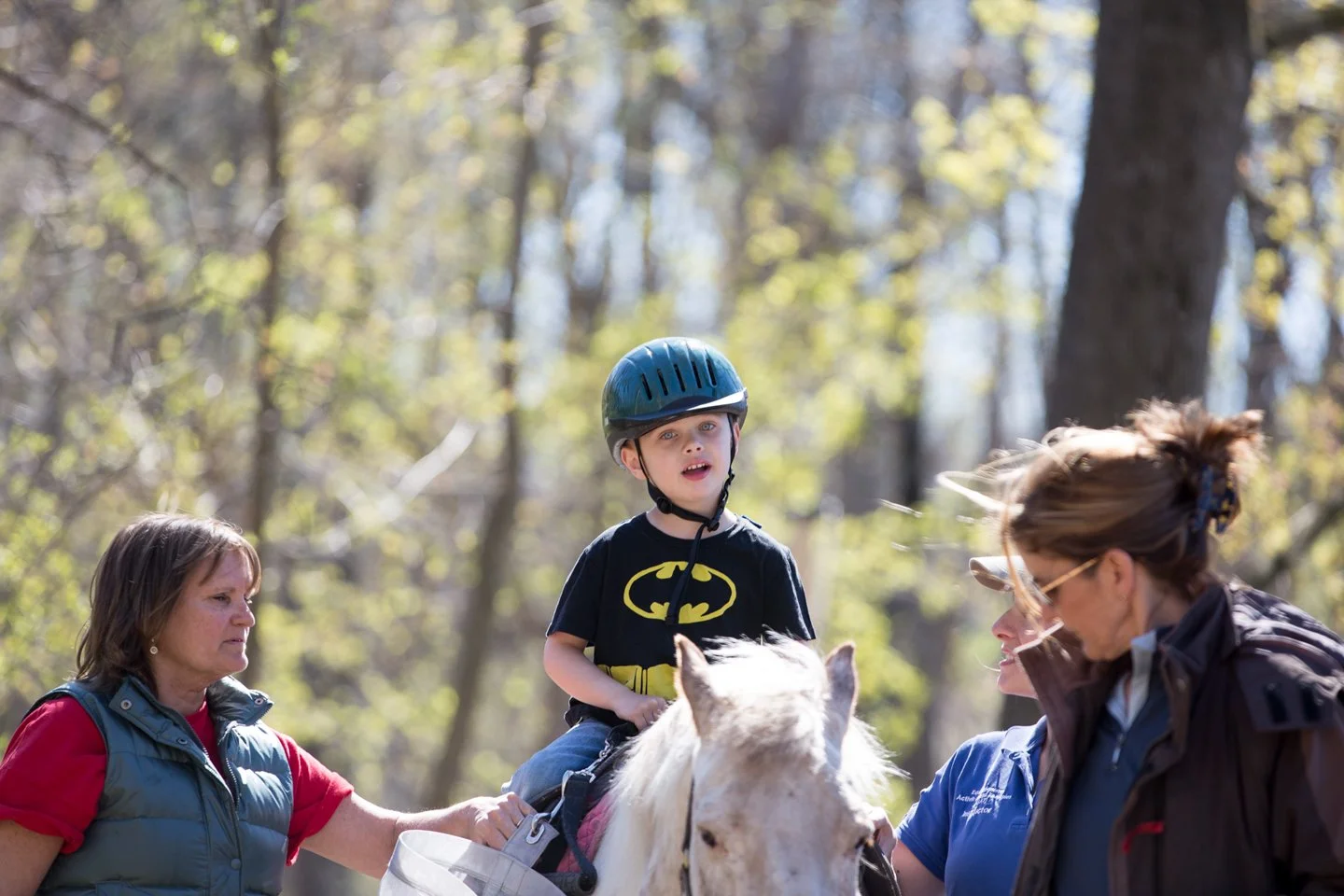 Young boy in a Batman t-shirt and safety helmet riding a white horse in a wooded area, with two women guiding and talking to him.