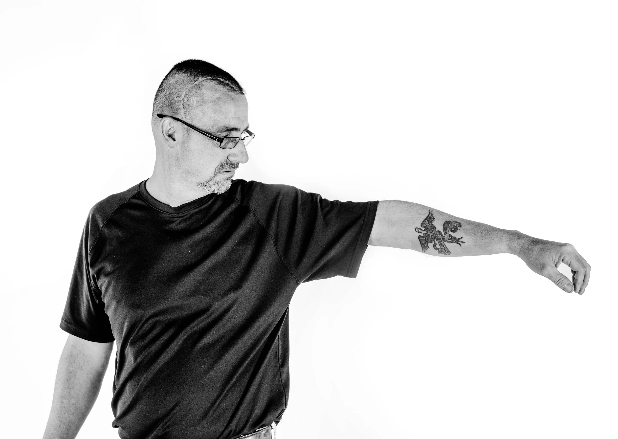 A man wearing glasses and a black t-shirt extends his arm to the side, showing a tattoo of a dragon and a sword on his forearm against a plain white background.