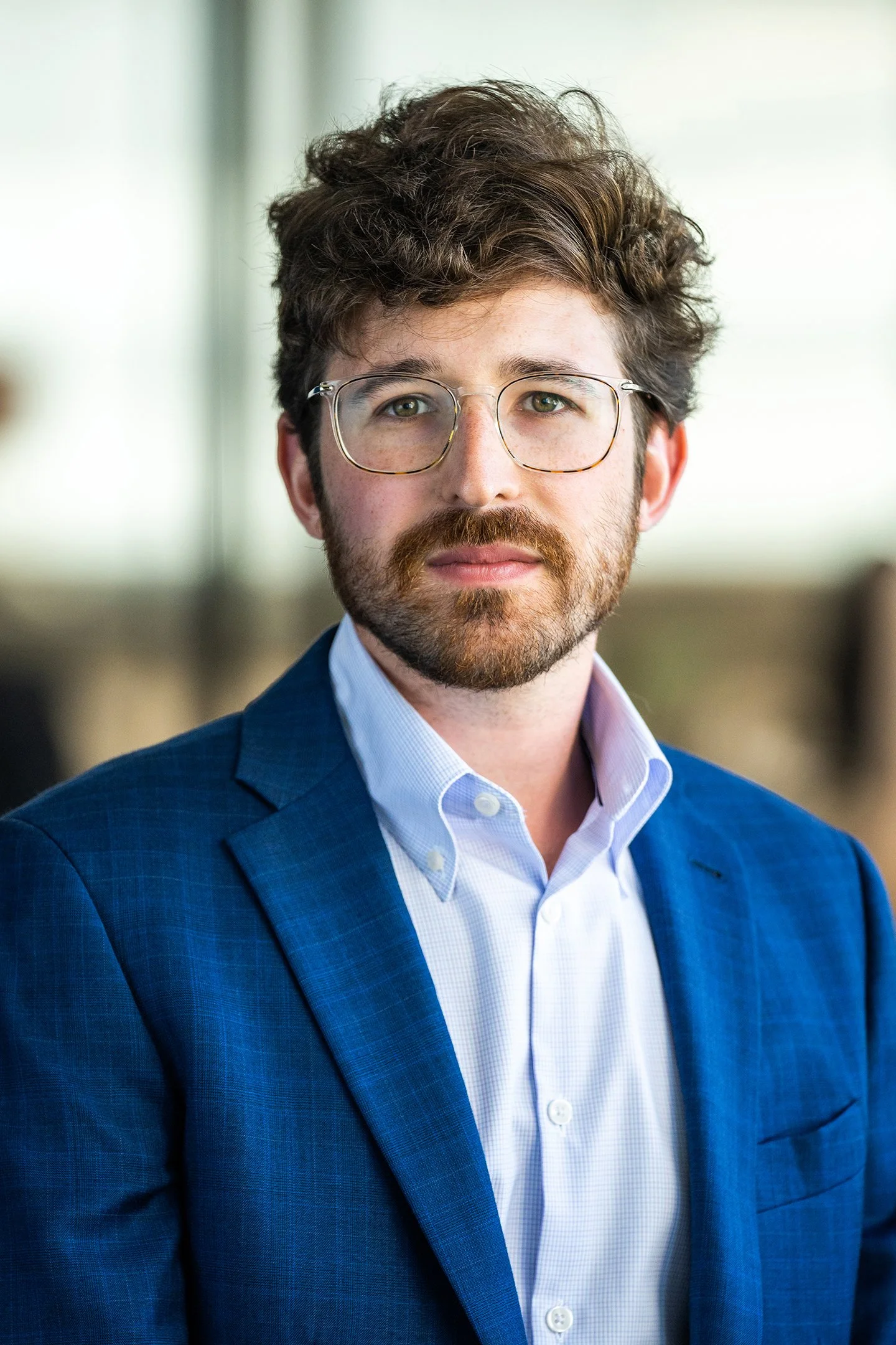 A young man with curly brown hair, wearing glasses, a blue suit, and a light blue shirt, looking directly at the camera.