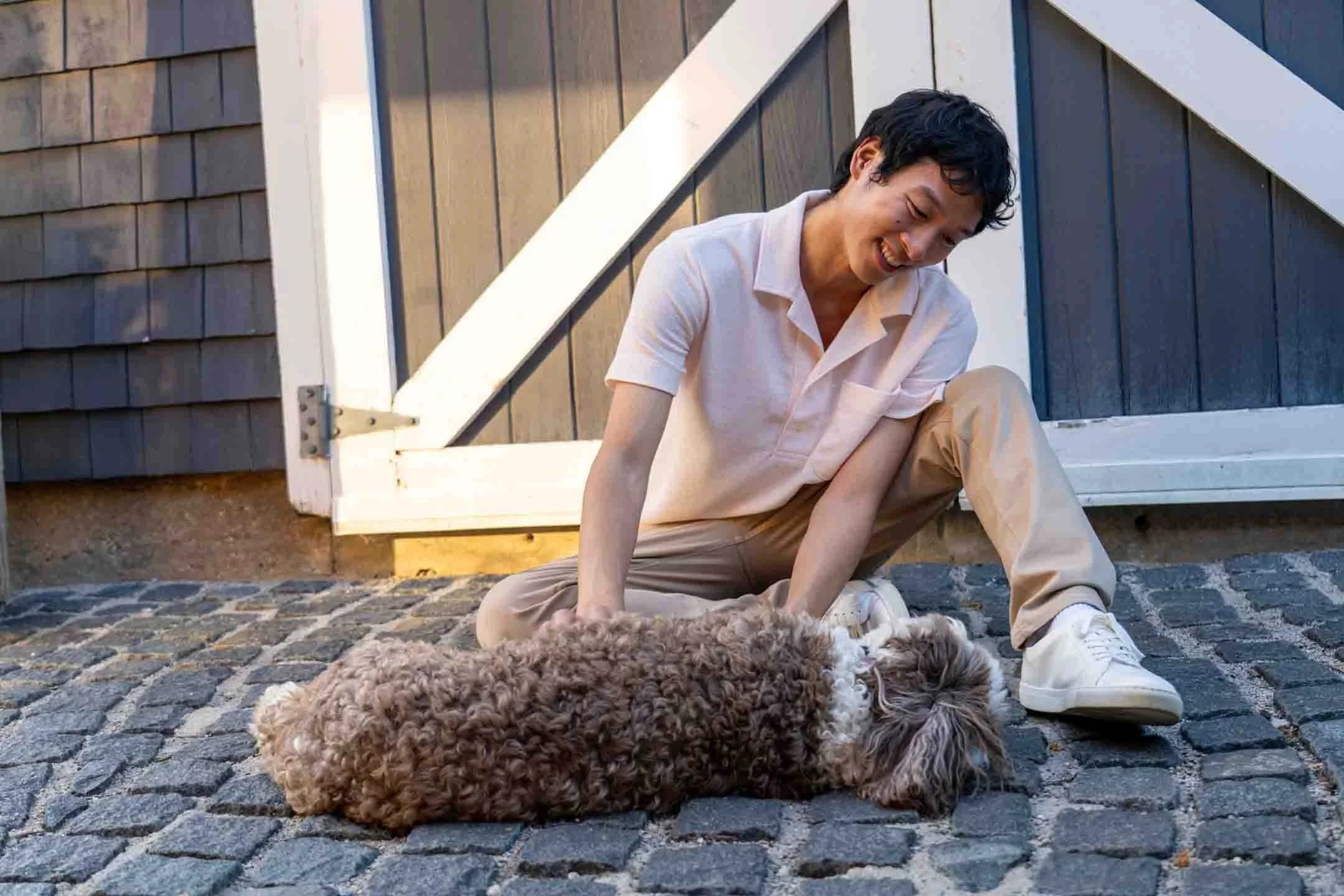 A young man with dark hair sitting on cobblestone ground, smiling and playing with a small curly-haired dog lying on its back, outside near a rustic building with dark wooden siding and white trim.