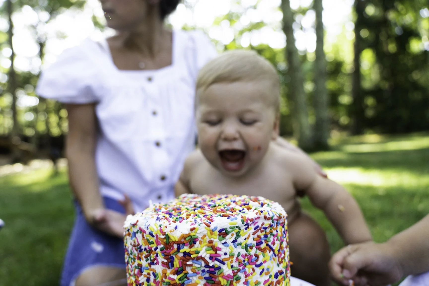 A young child with blond hair celebrating a birthday outdoors, leaning towards a large, colorful cake with rainbow sprinkles and white frosting, with a woman in a white top standing behind him.