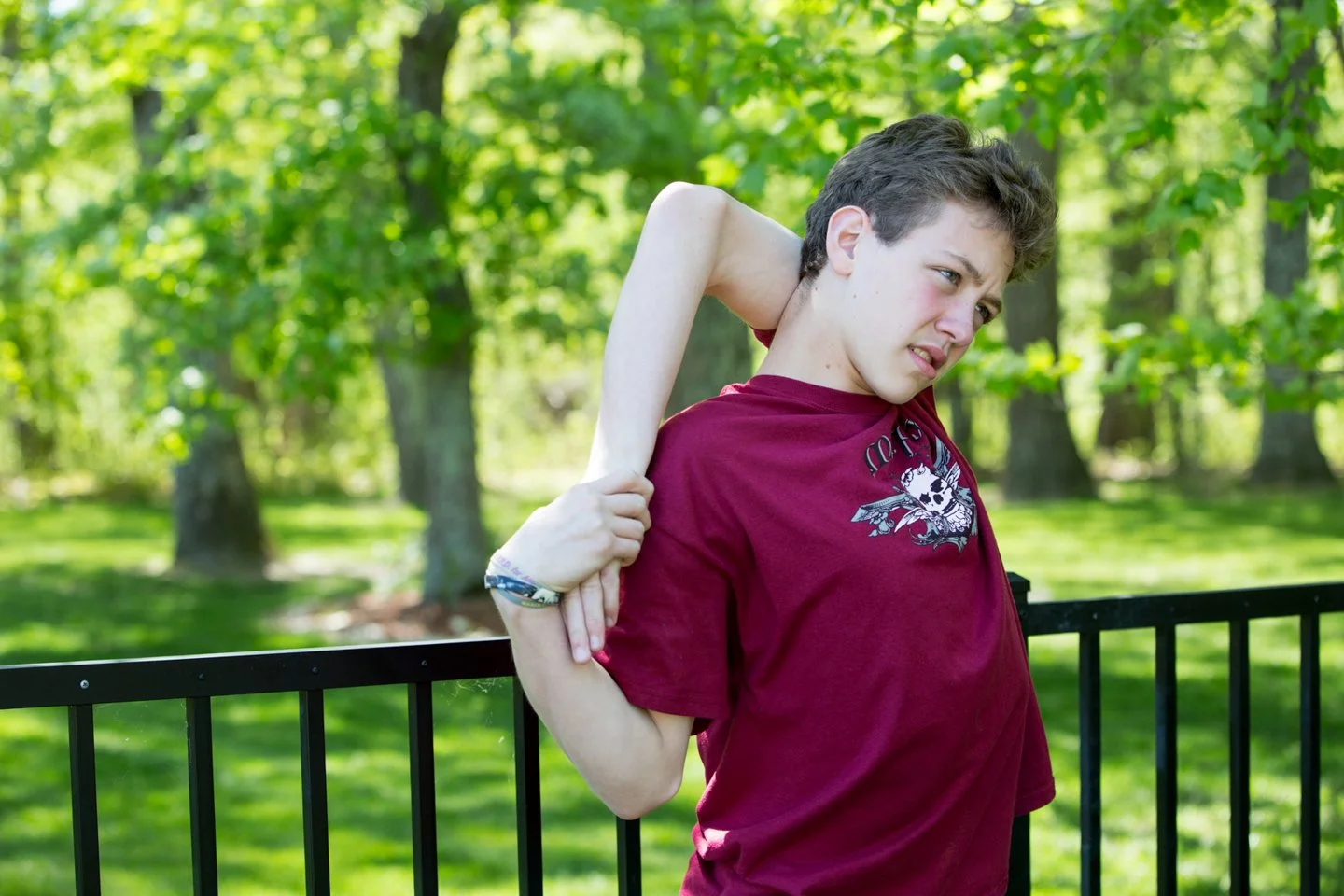 A teenage boy stretching outdoors in a park with trees and grass, wearing a maroon T-shirt with a graphic design.