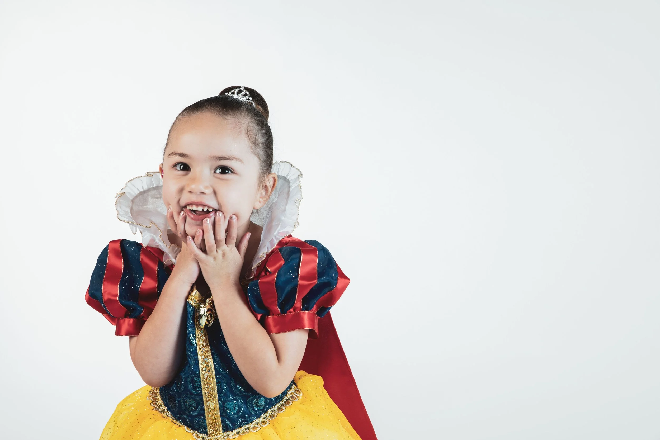 Young girl wearing a Snow White costume with a yellow skirt, blue top, and red cape, holding her face with a joyful expression.