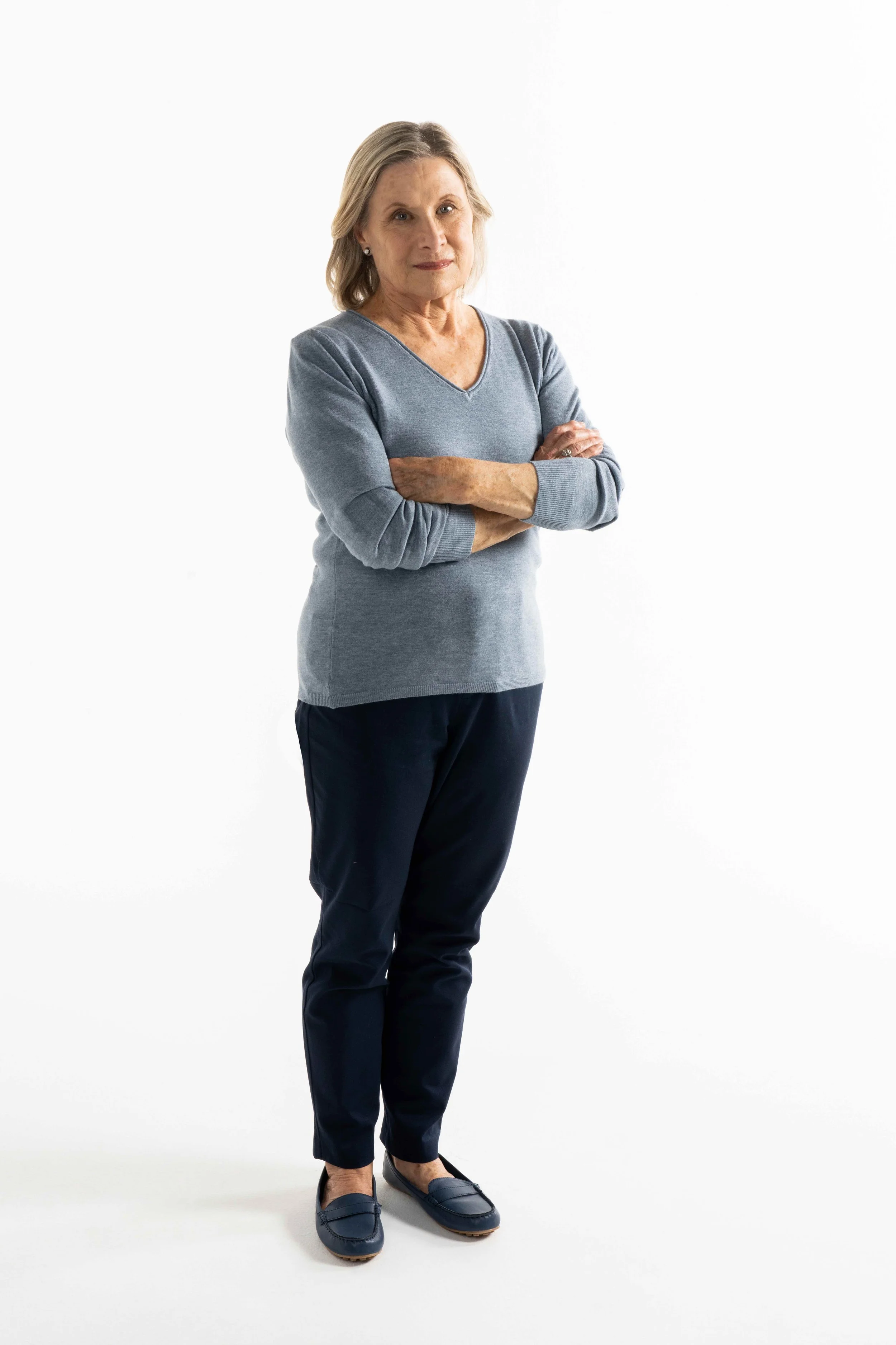 An older woman with gray hair, wearing a gray long-sleeve shirt and navy pants, standing with her arms crossed against a plain white background.