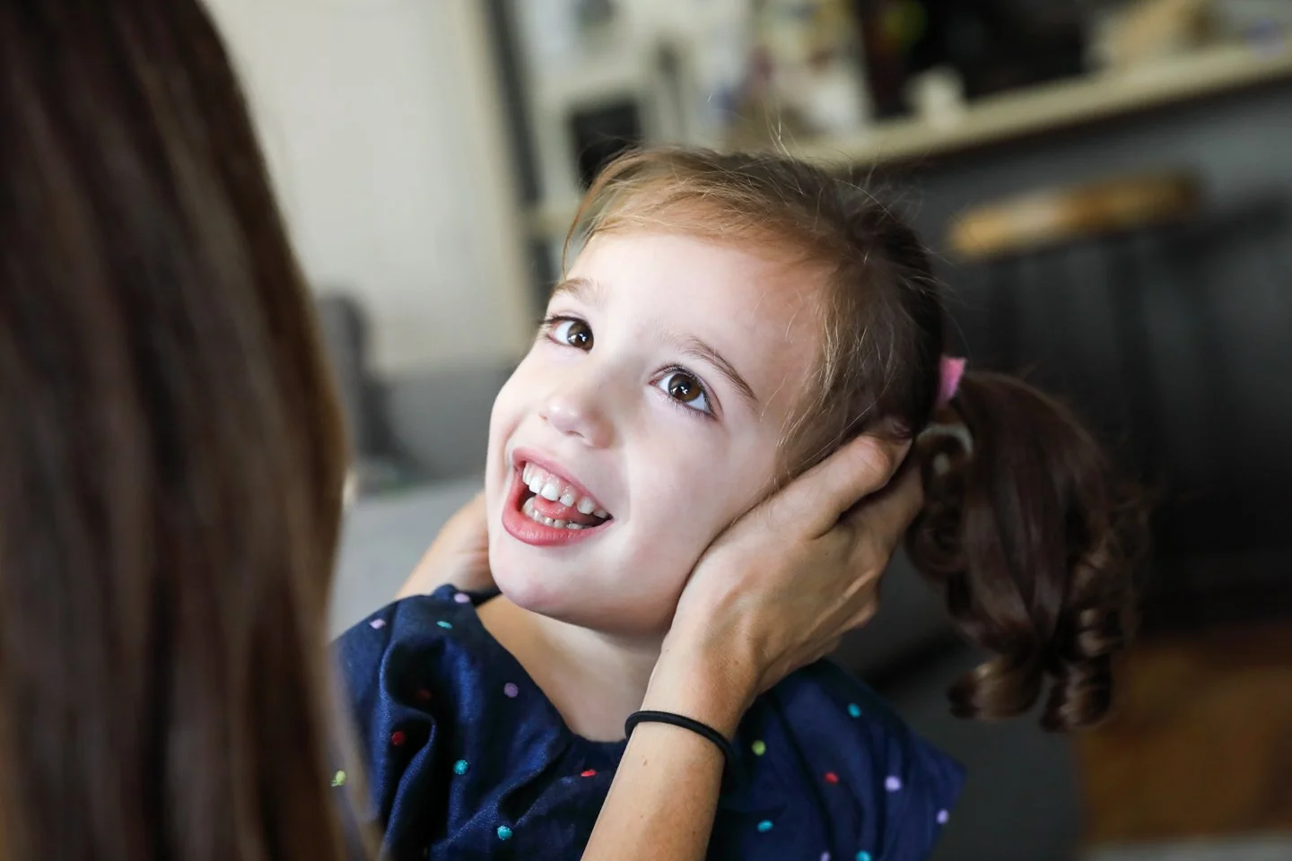A young girl with brown hair in pigtails and a blue shirt with multicolored dots smiles while an adult gently holds her face and smiles in a cozy living room.