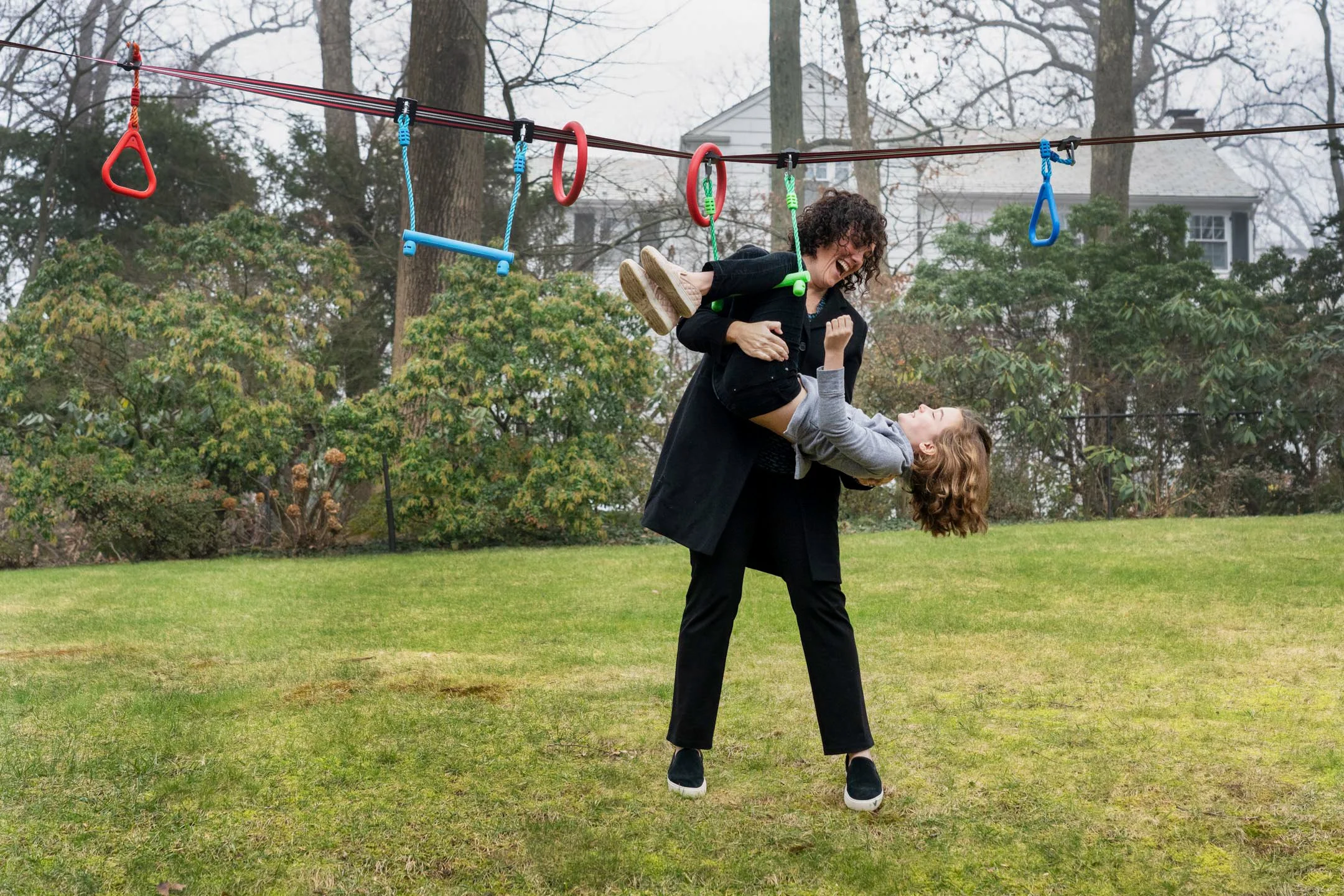 Two women playing on a backyard playground; one woman is lifting the other woman, who is laughing and lying back in her arms, while they are on grass with trees and houses in the background.