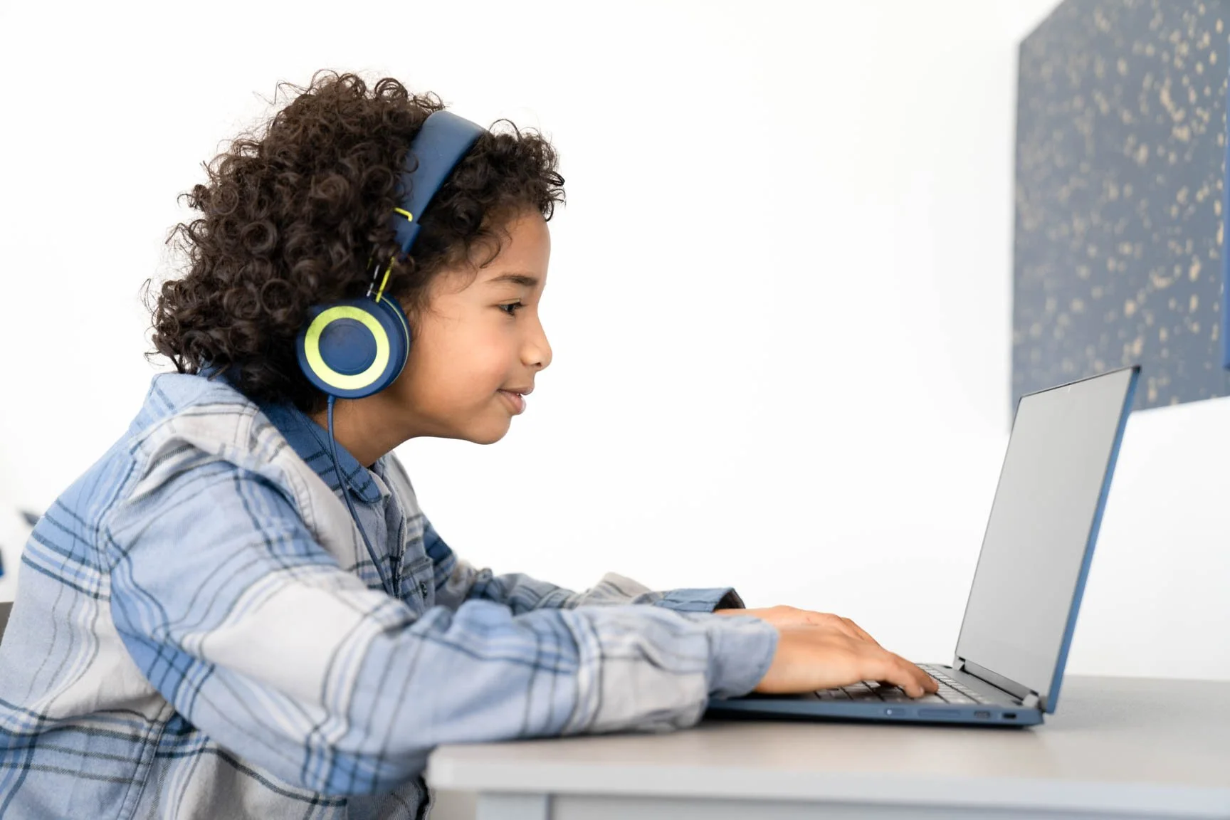 Young boy with curly hair wearing blue headphones using a laptop at a desk.