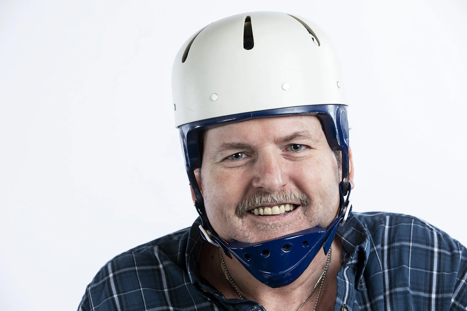 A man smiling wearing a white and blue helmet, dressed in a checkered shirt against a plain white background.