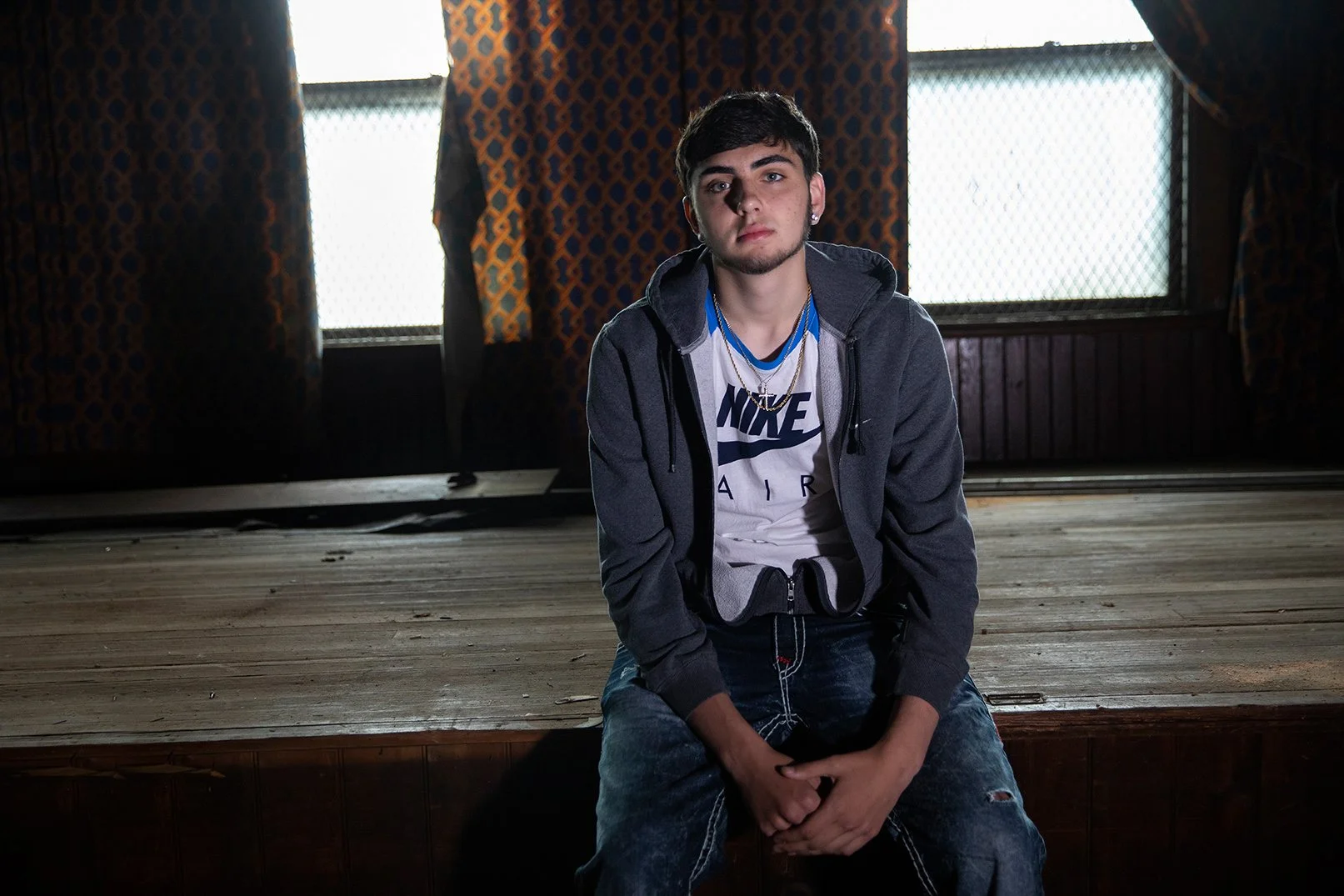 Young man sitting on the floor in front of a stage with curtains and windows in the background, wearing a Nike shirt, gray hoodie, and blue jeans.