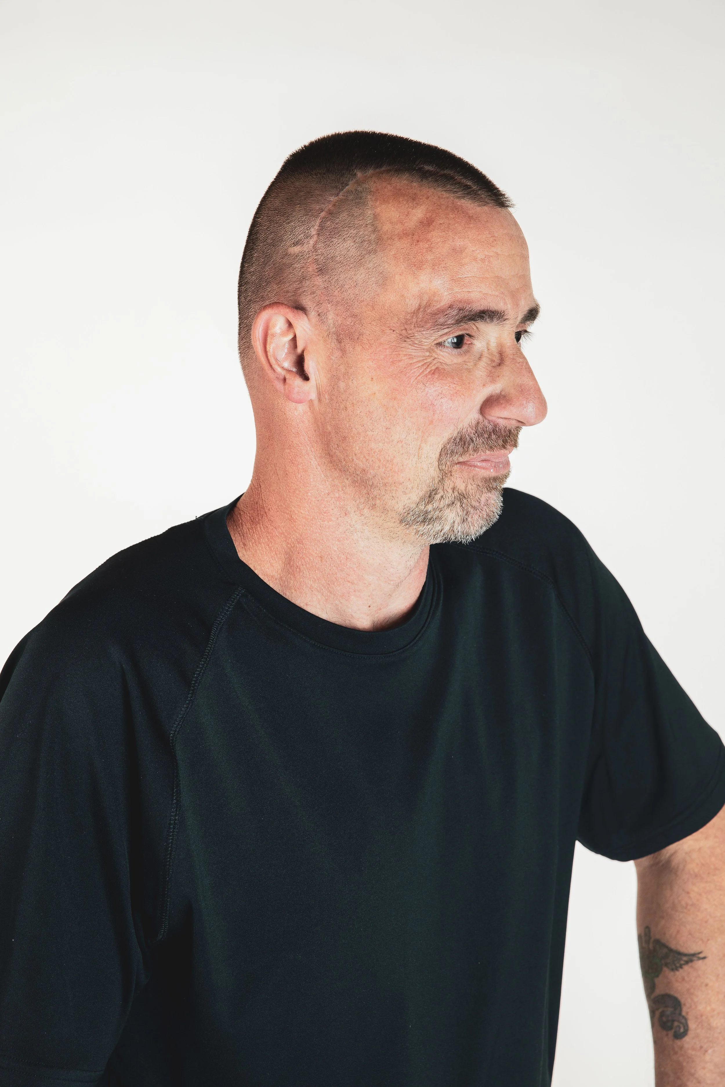 Side profile of a man with a buzz cut and facial hair, wearing a black T-shirt, against a plain white background.