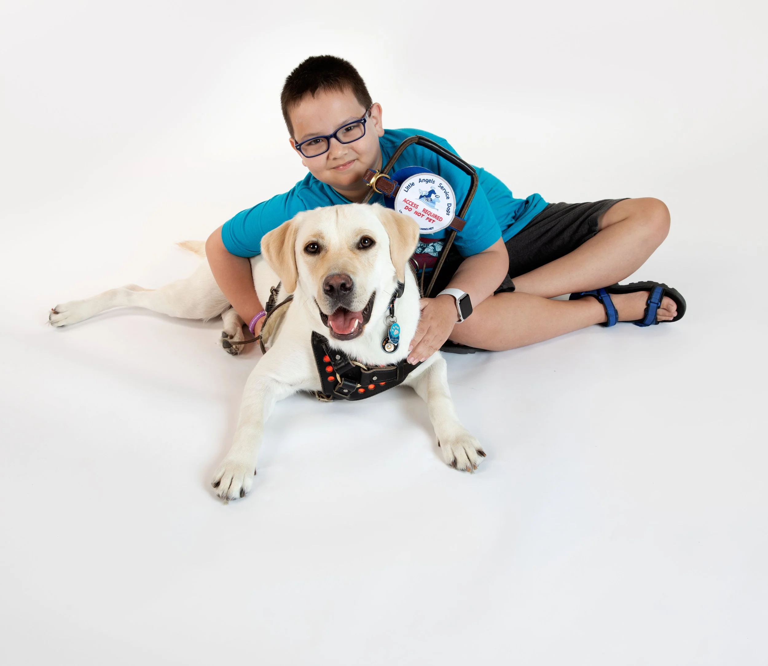 A young boy sitting on the floor with a service dog, both looking happy. The boy wears glasses and a blue shirt, the dog is wearing a harness and service tags.