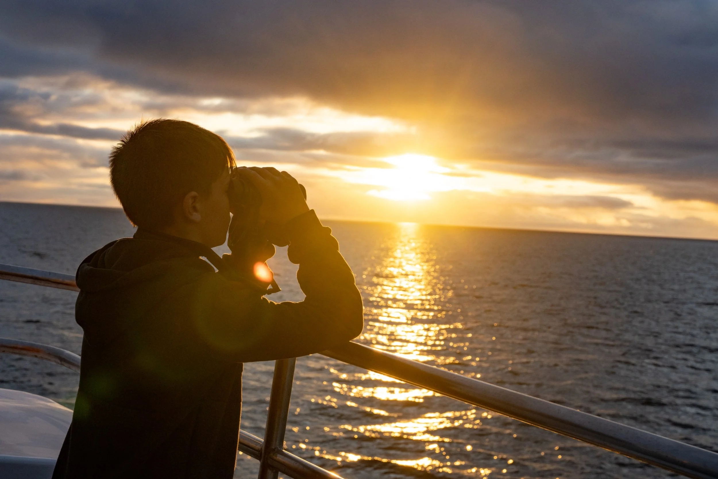 Person taking a photograph or looking through binoculars on a boat during sunset over the ocean.