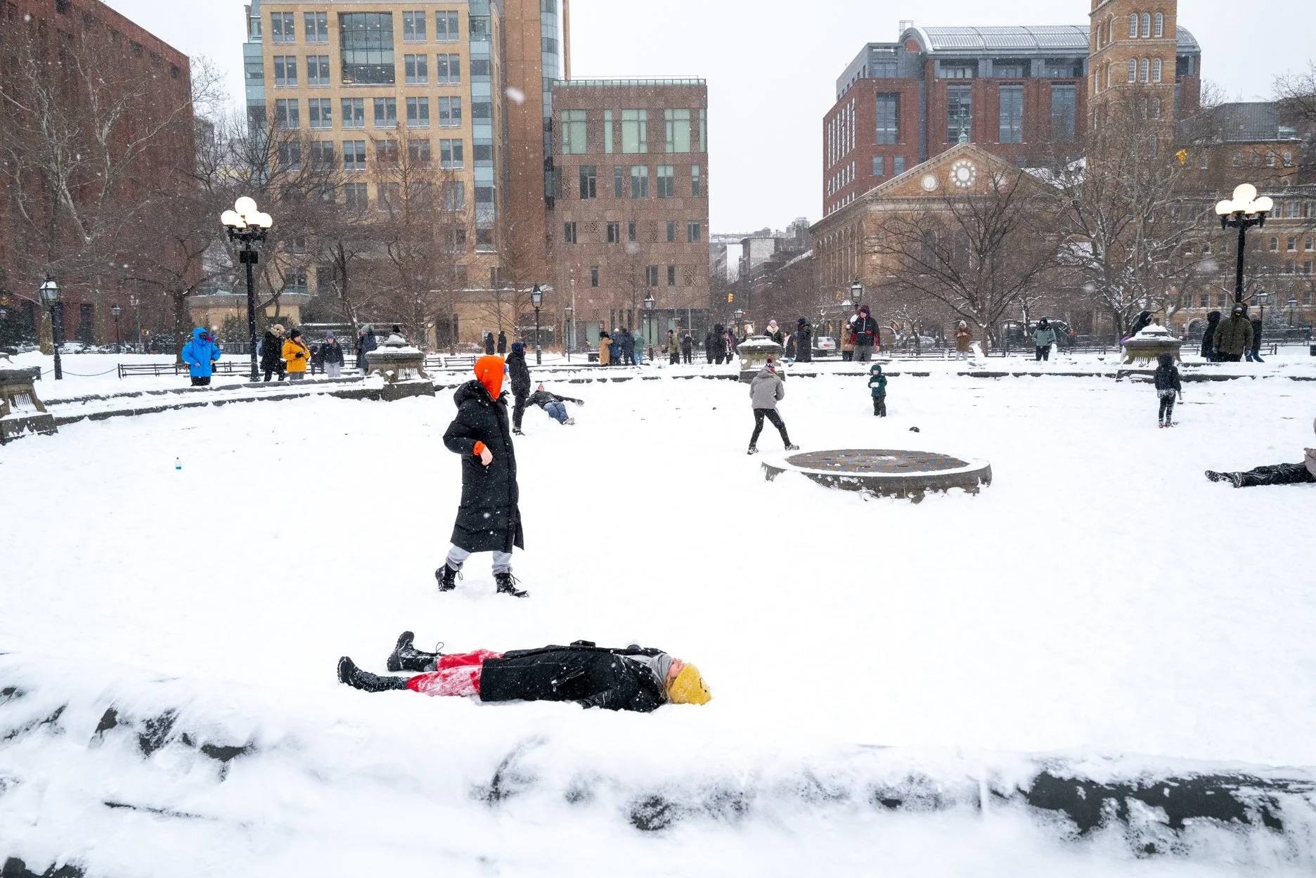 People playing in a snow-covered park in an urban area, some standing, some sitting, with a person lying on the snow in the foreground.