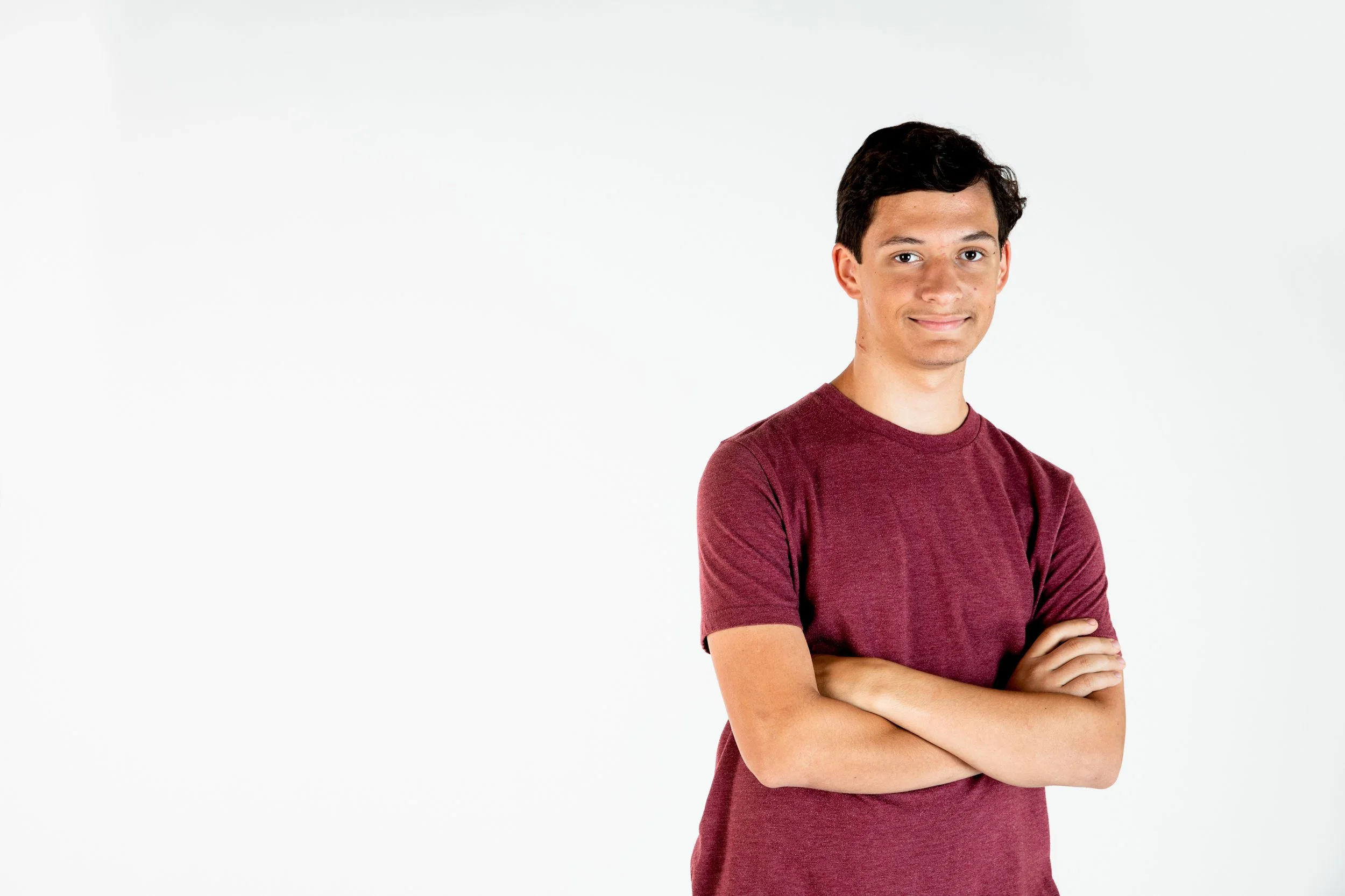 A young man with short dark hair, wearing a maroon t-shirt, standing with arms crossed and smiling against a plain white background.