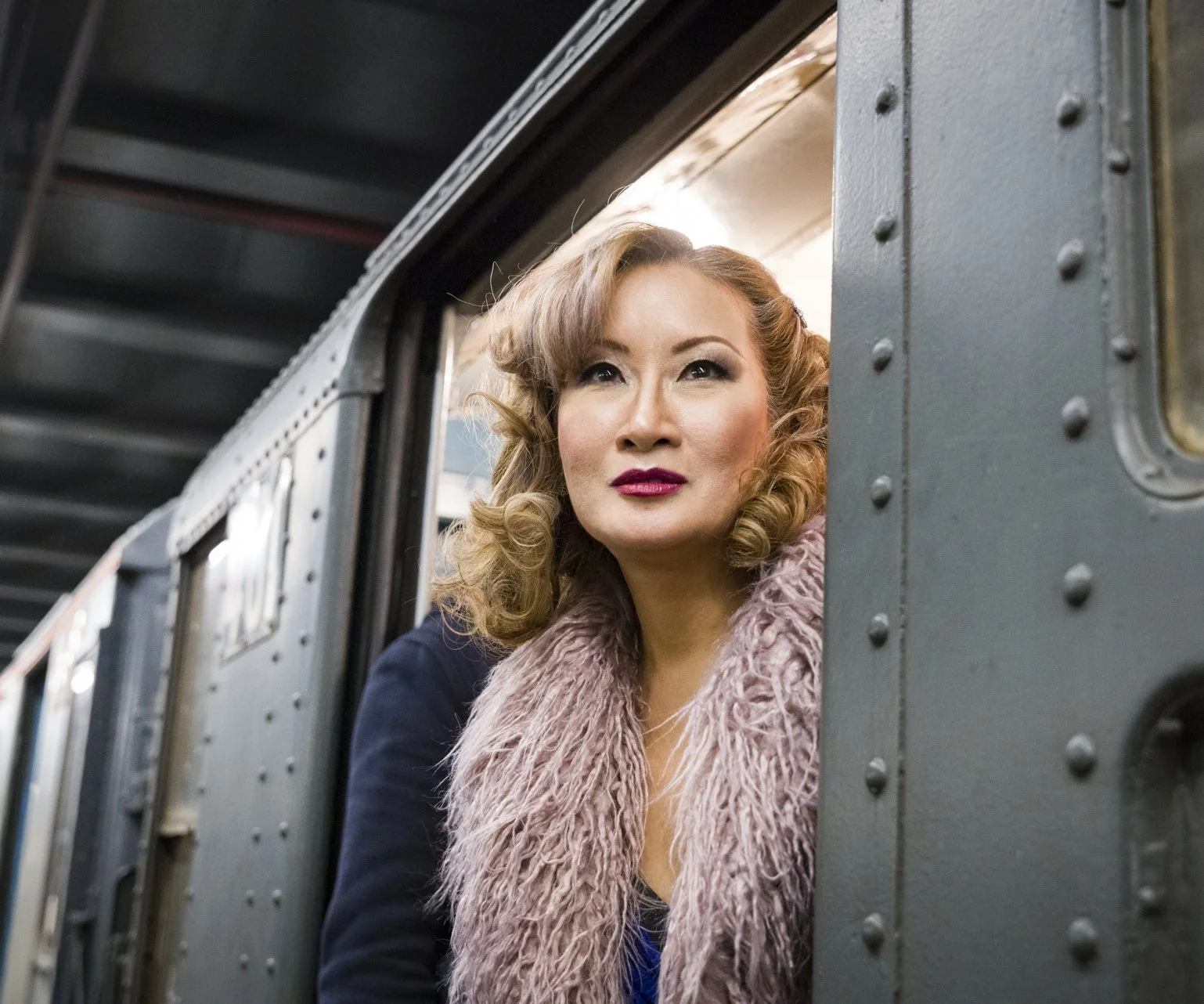 Woman with blonde curly hair wearing a fur-like scarf and dark jacket looking out of train window.