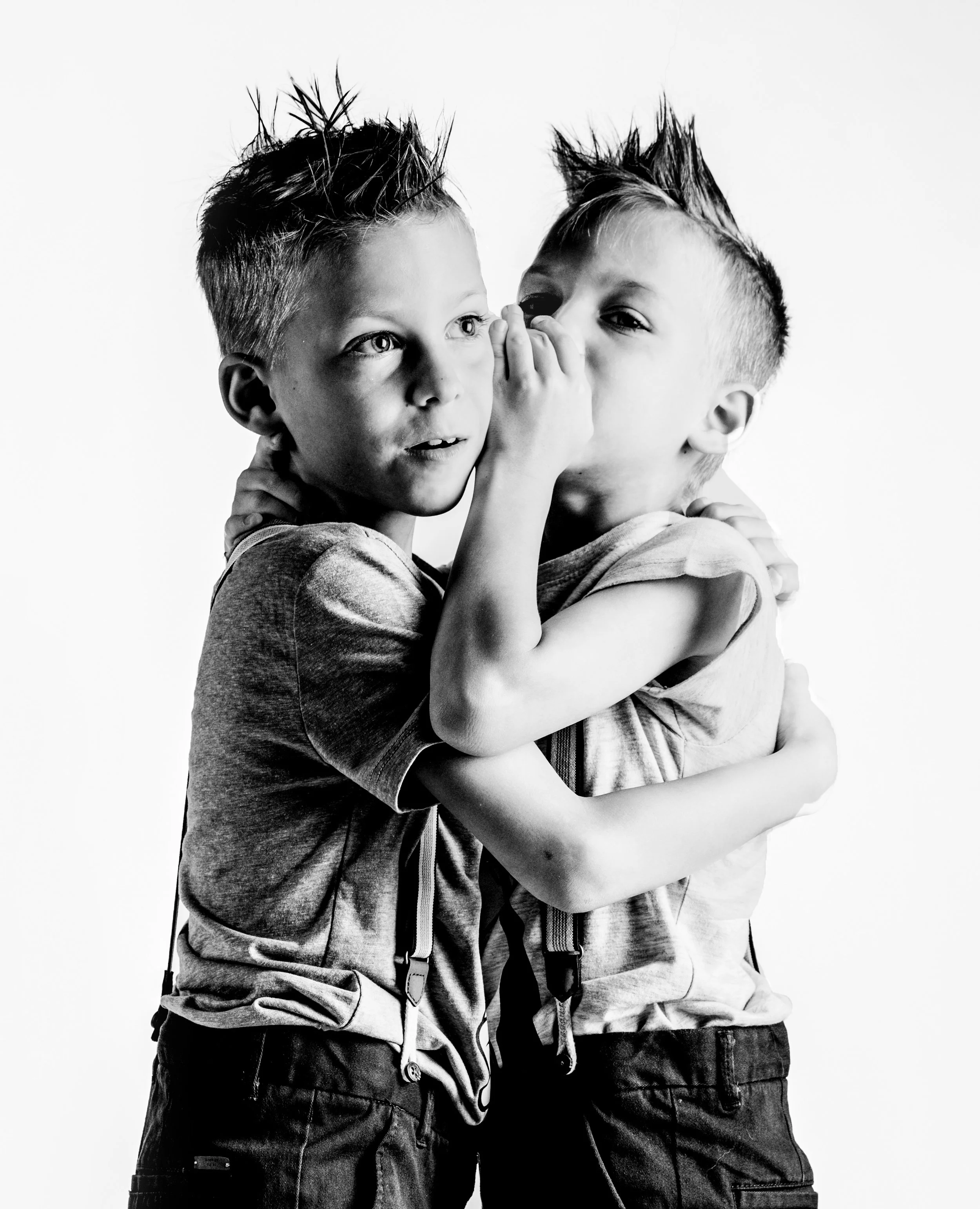 Two young boys embracing, one whispering into the other's ear, in a black and white photograph.
