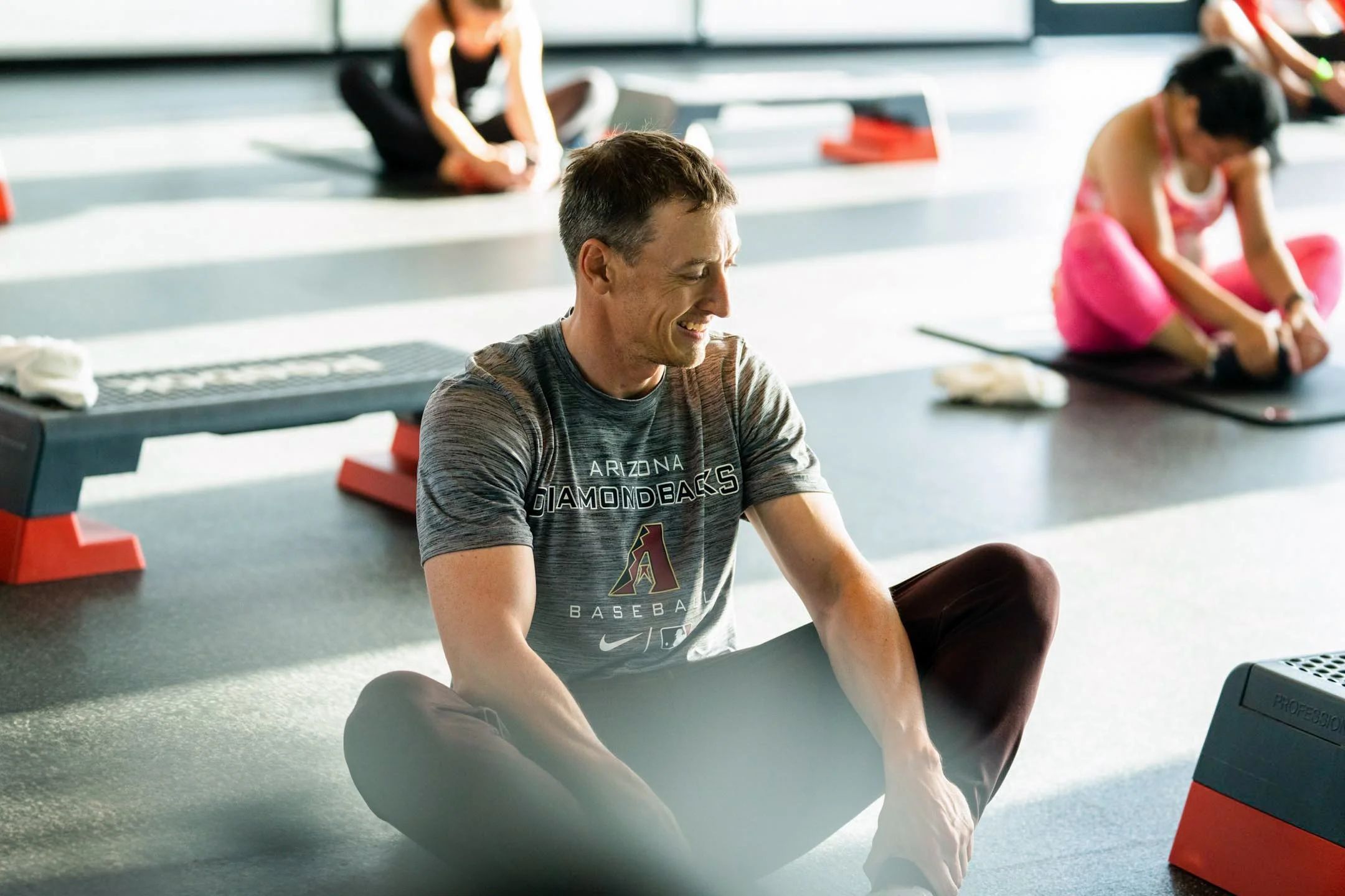 Man in gray Arizona Diamondbacks t-shirt sitting cross-legged on indoor gym floor, smiling, with women in workout attire stretching in the background.