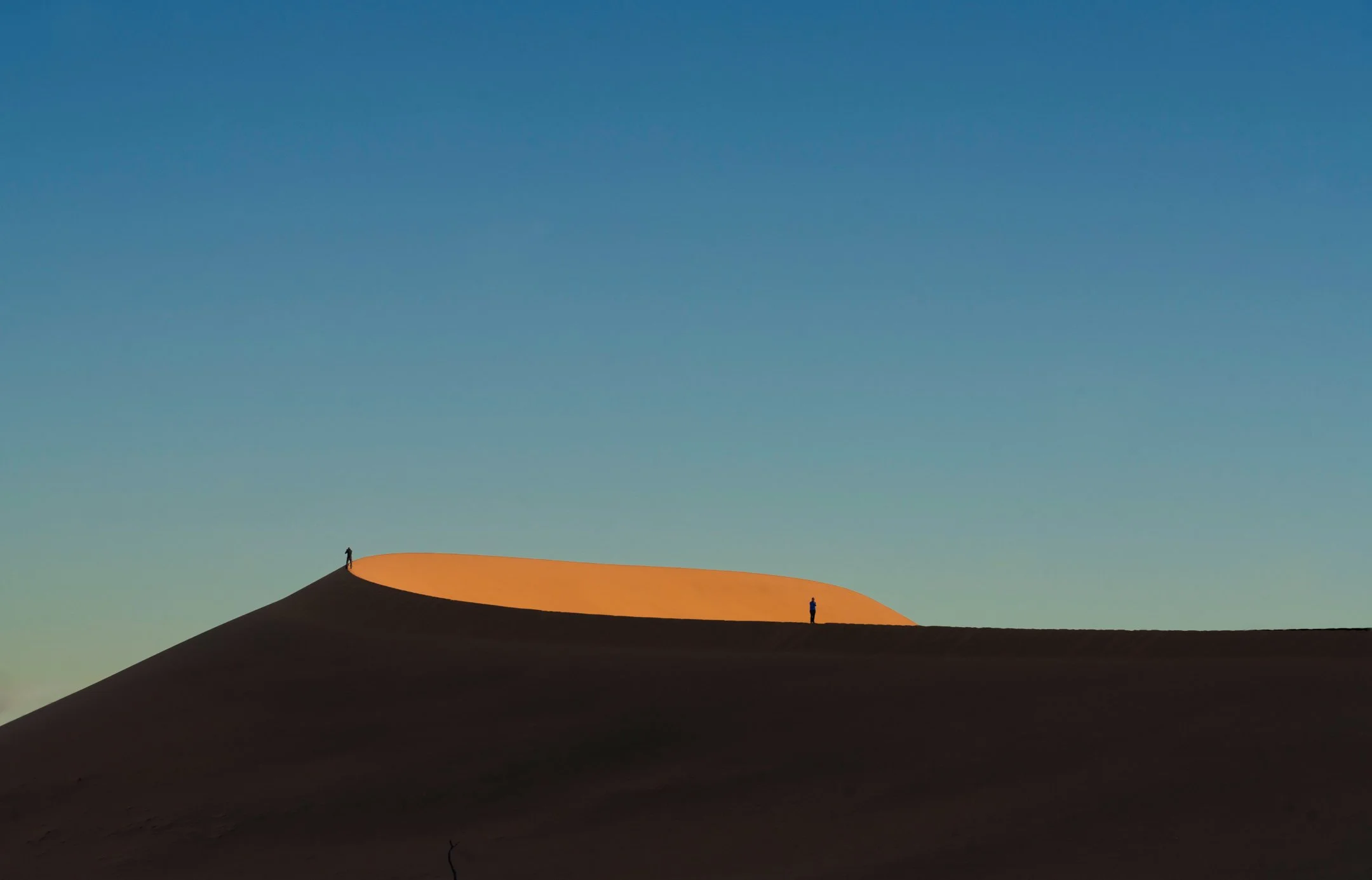 Two small figures standing on sand dunes against a clear blue sky, with the dunes casting shadows.