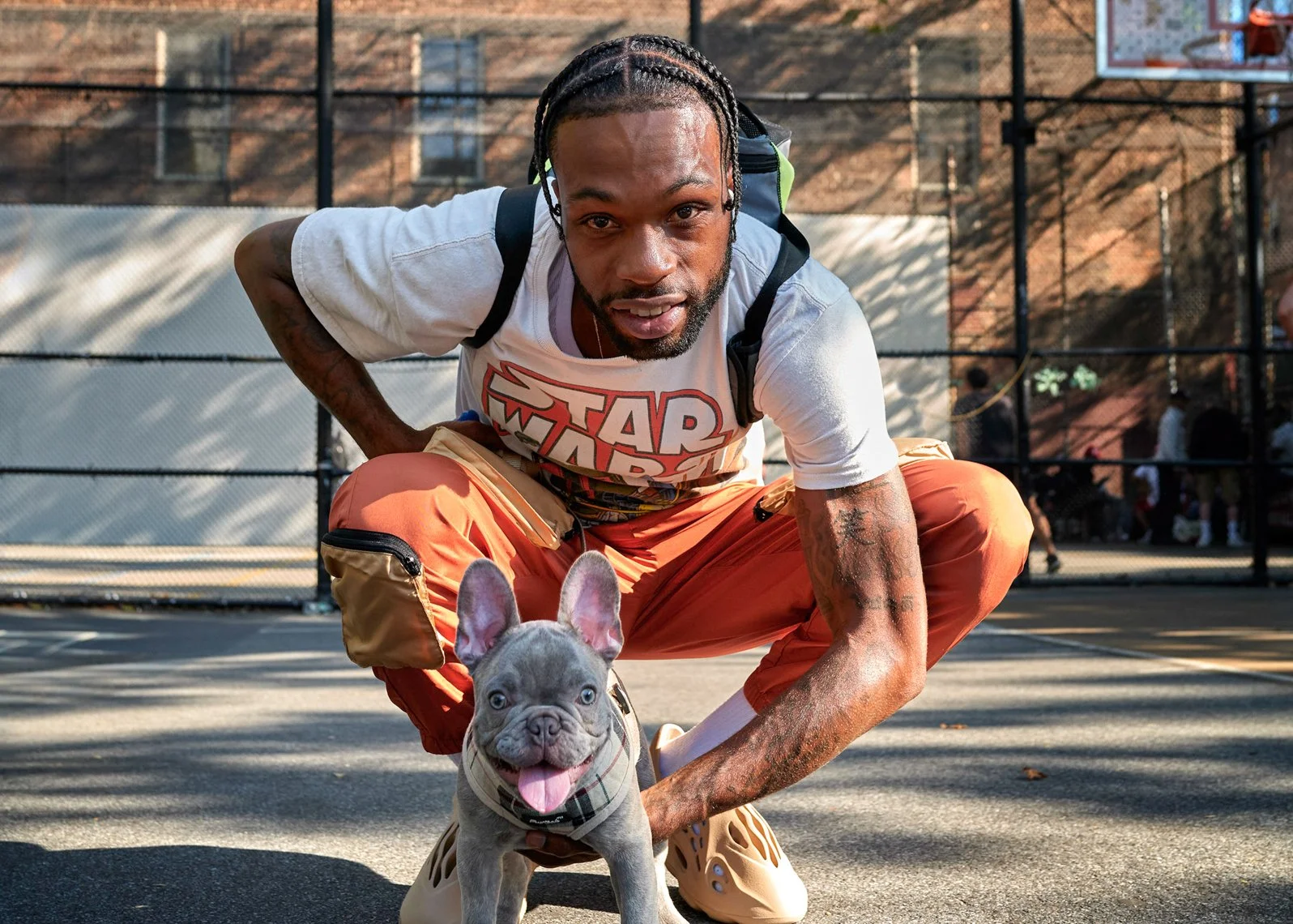 A man with braided hair crouches down next to a gray French Bulldog puppy on a basketball court, with a chain-link fence and other people in the background under a sunny sky.