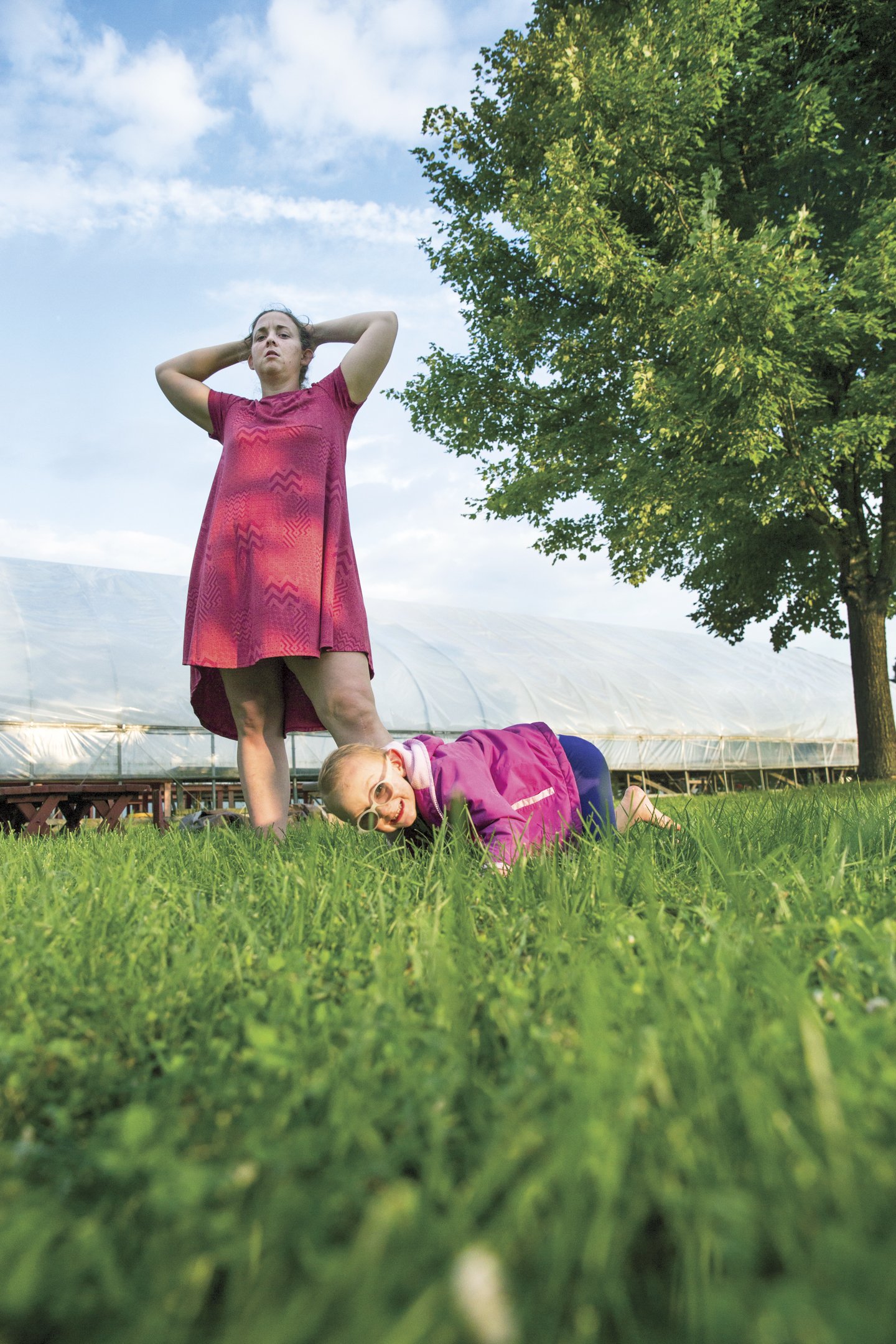 Two young women outdoors on grass, one standing with hands behind head and the other crawling on all fours, wearing casual clothes and sunglasses, under a blue sky with trees and a greenhouse in the background.
