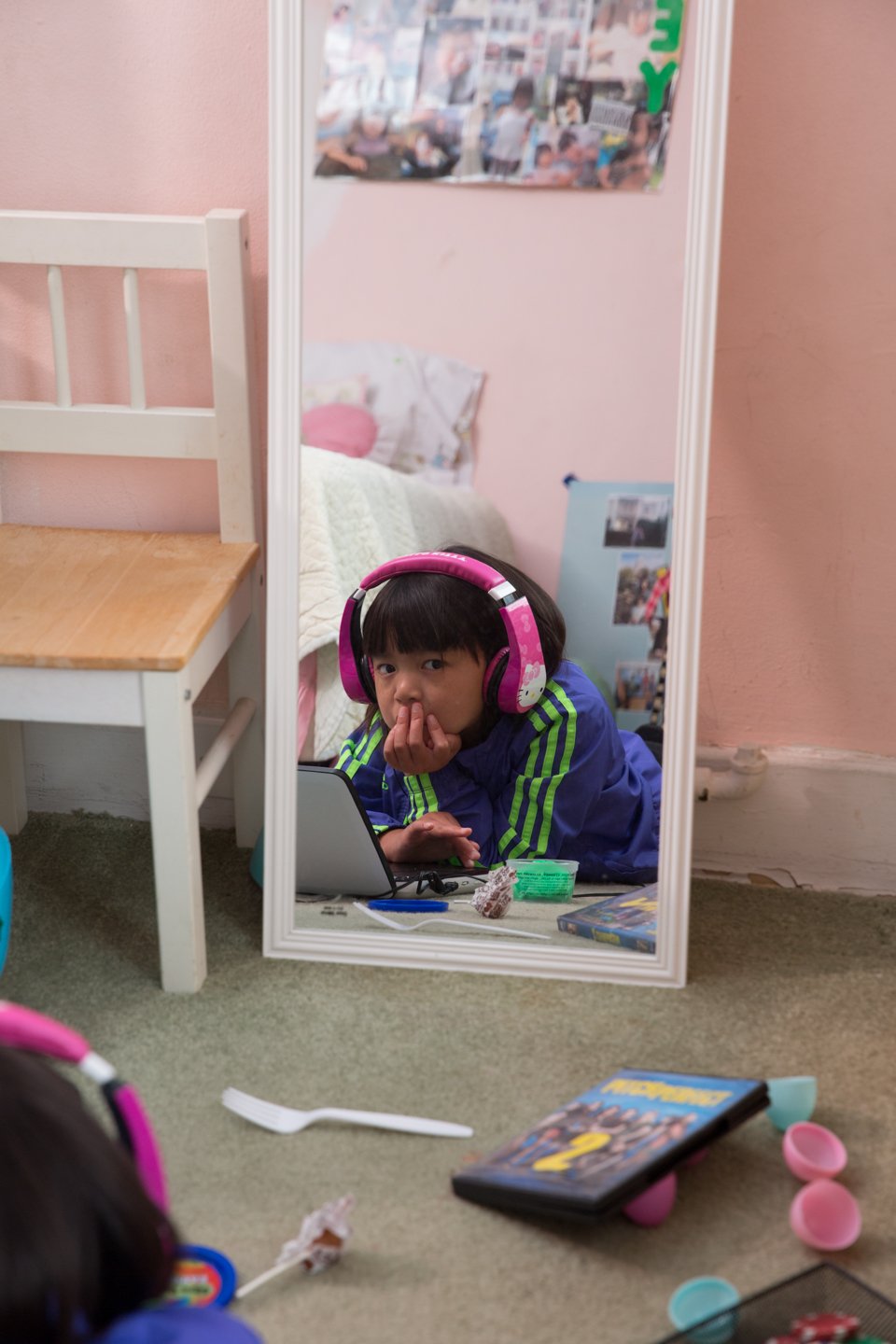 Child with black hair wearing pink headphones, looking into a mirror on the floor, with toys and a book nearby.