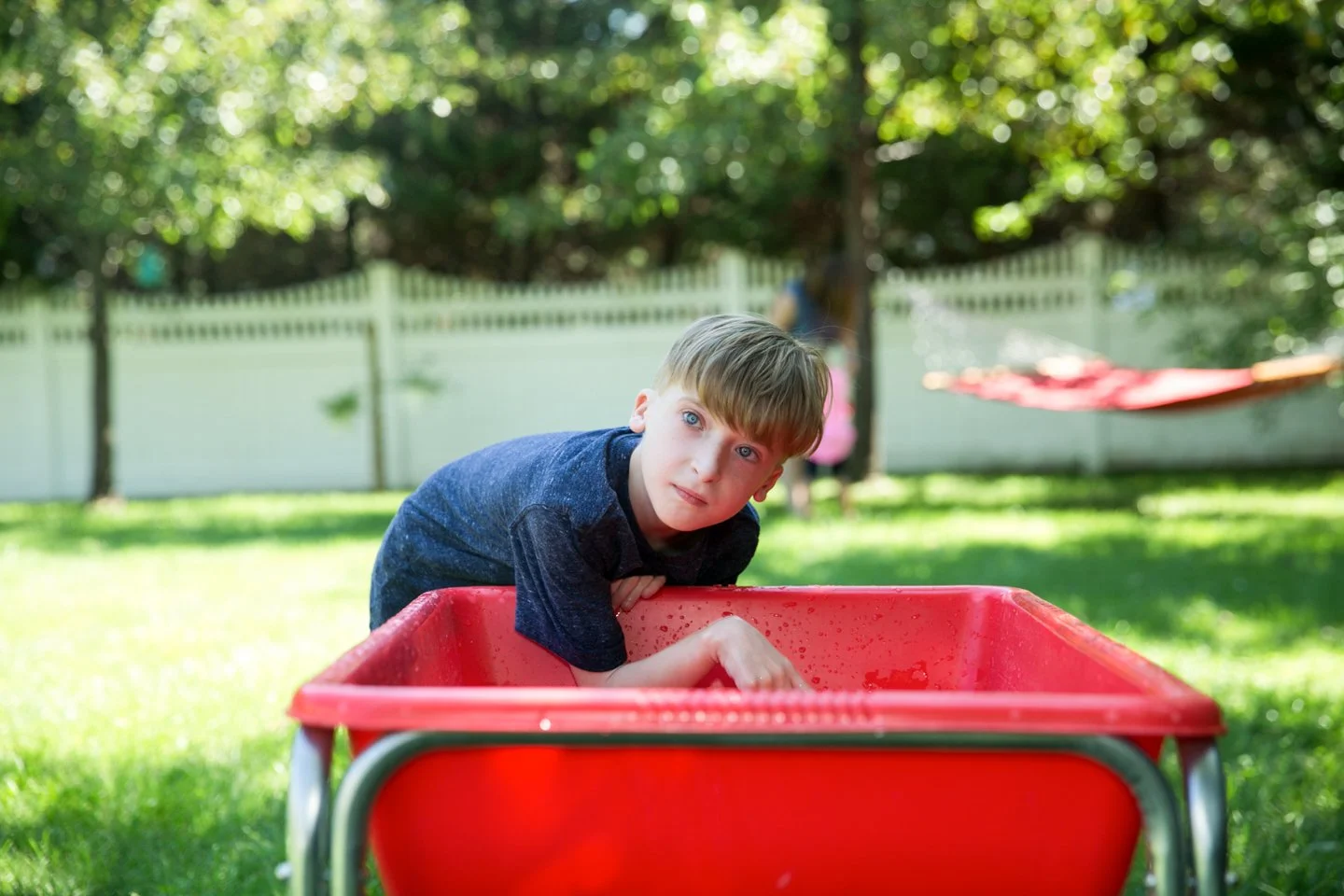 A young boy leaning over a red plastic wagon in a grassy backyard, with trees, a white fence, and a clothesline with a red sheet in the background.