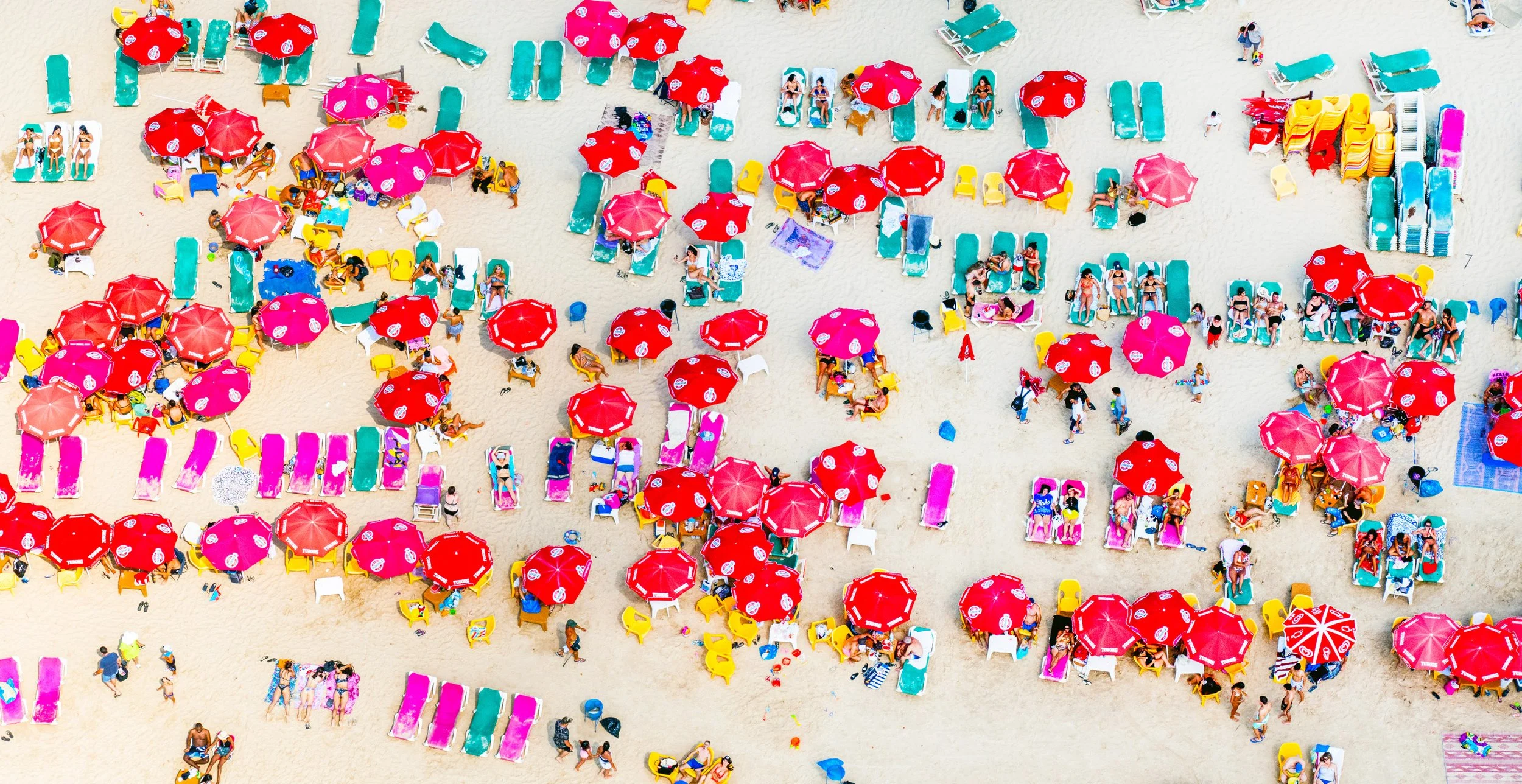 Aerial view of a crowded beach with colorful umbrellas, lounge chairs, and people sunbathing and walking on the sand.