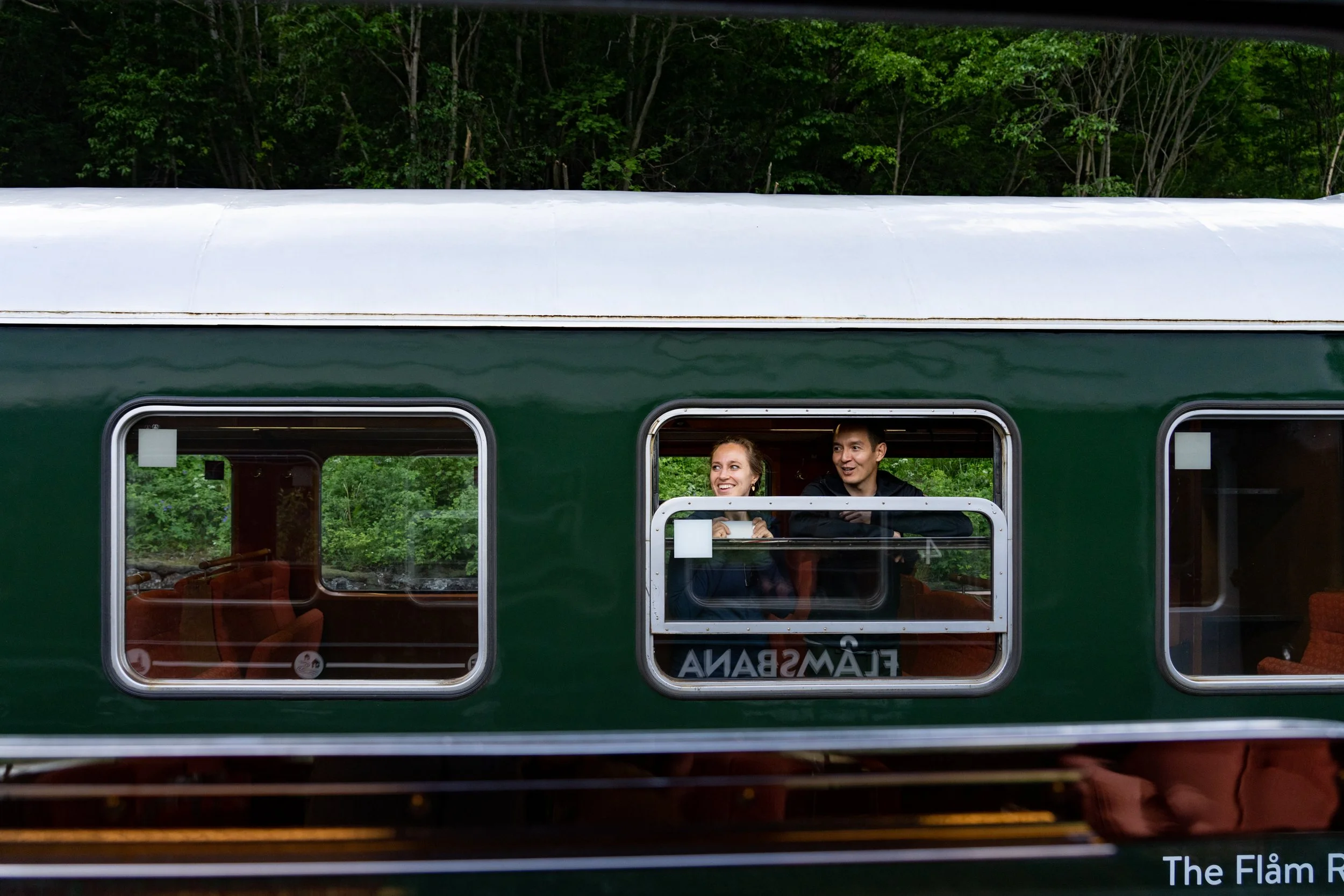 Two people smiling and looking out of a window of a green train with a forest background.