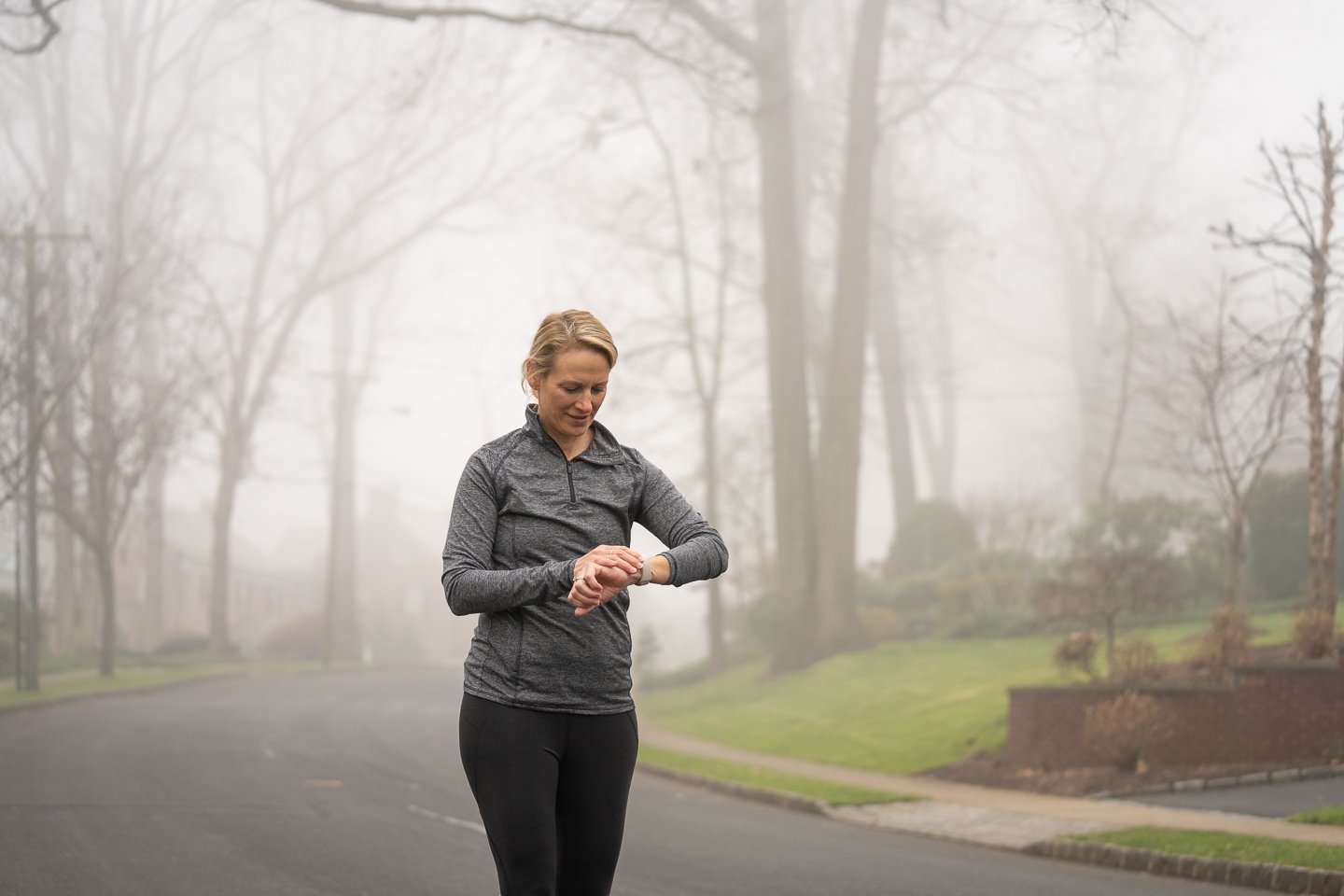 A woman in a gray athletic jacket and black leggings stands on a foggy outdoor path, looking at her smartwatch.
