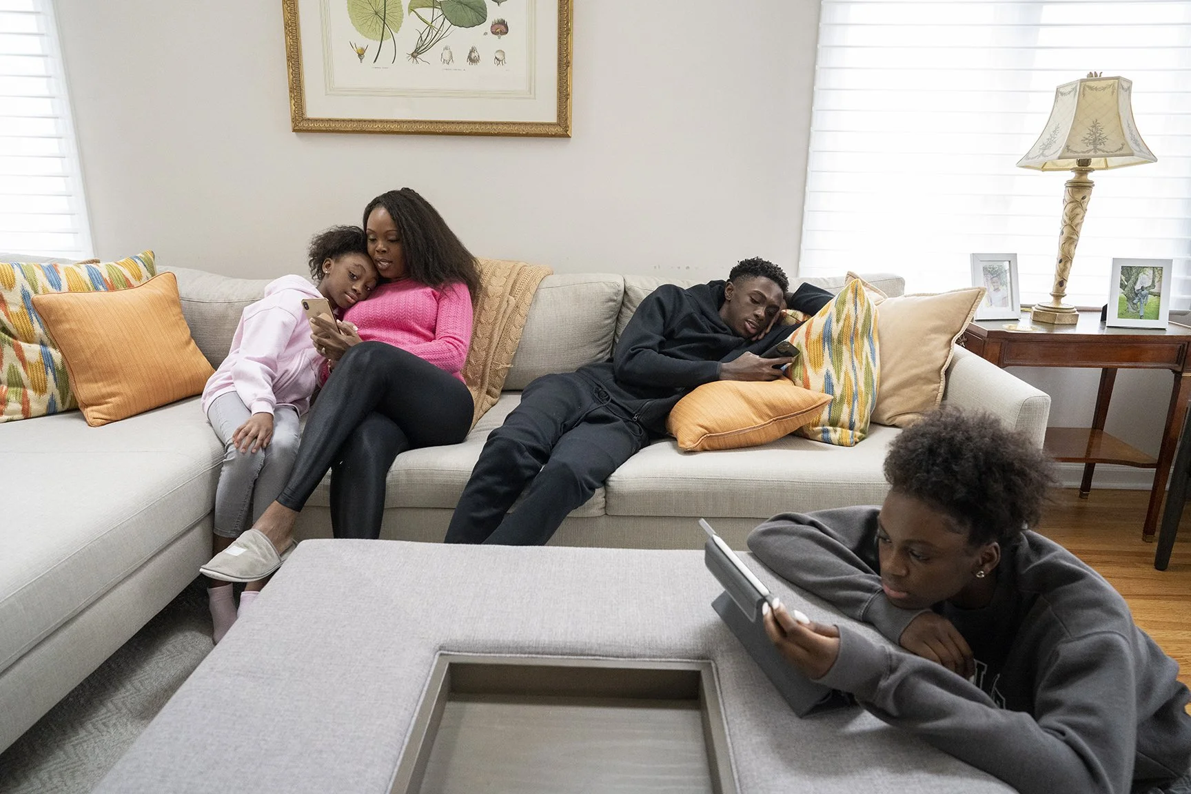Four children sitting and lying on a beige couch in a living room, using electronic devices like smartphones and tablets, with pillows, a lamp, and framed photos on a side table in the background.