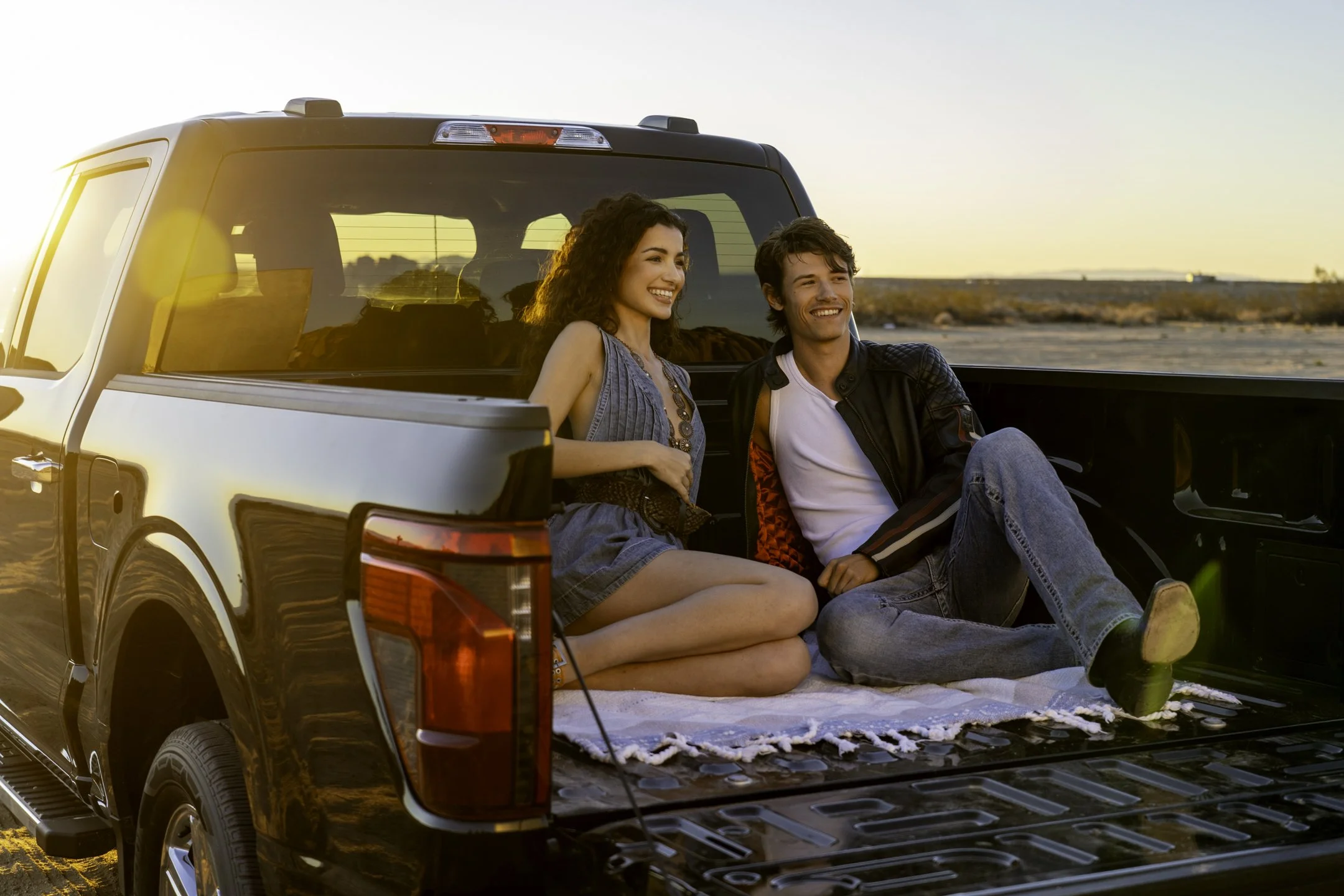 A young woman and man sitting in the cargo bed of a black pickup truck during sunset, smiling and enjoying the outdoors.