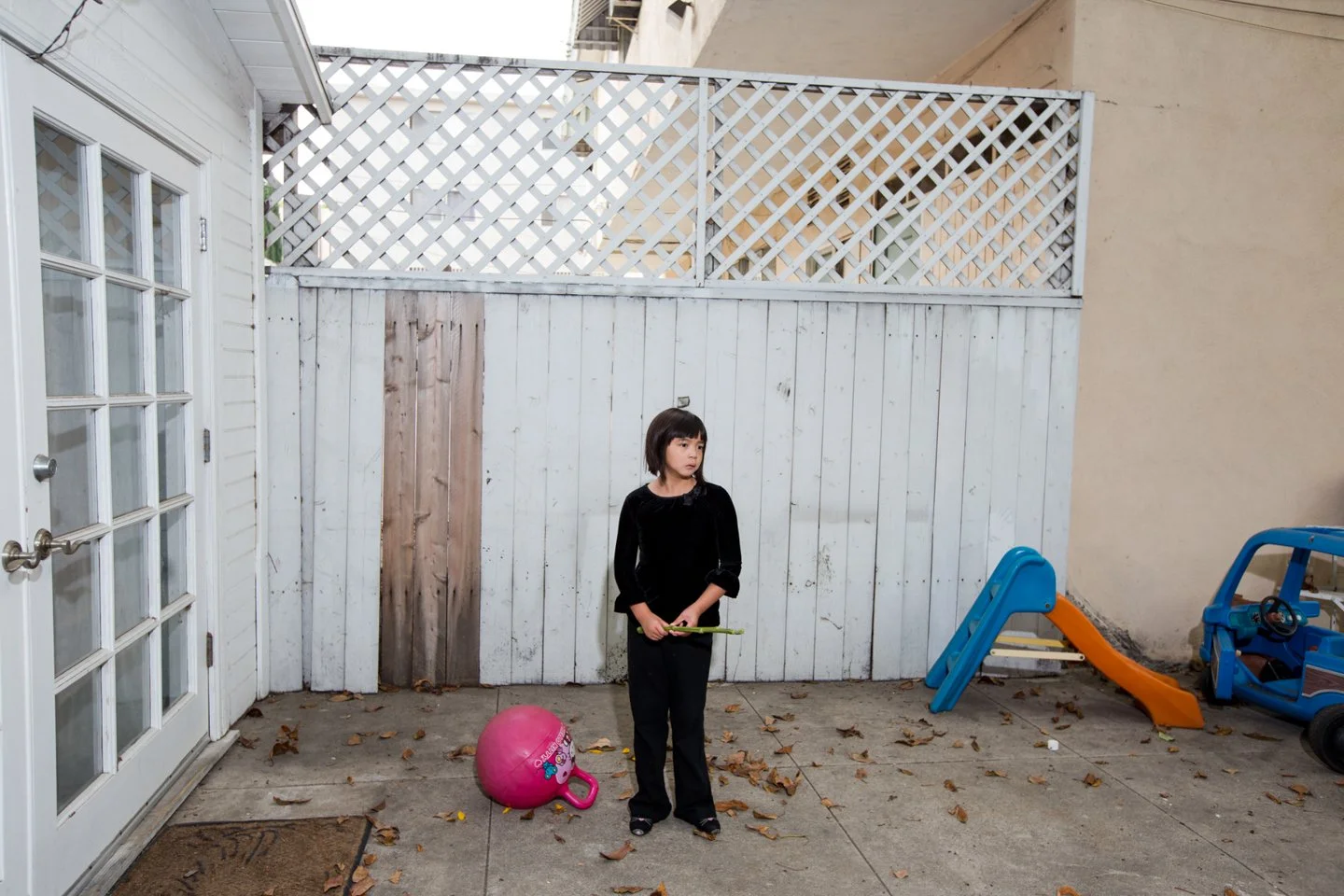 A young girl standing outside in a backyard with a pink ball, a toy slide, and a toy car. She is holding a green object and looking to the side.