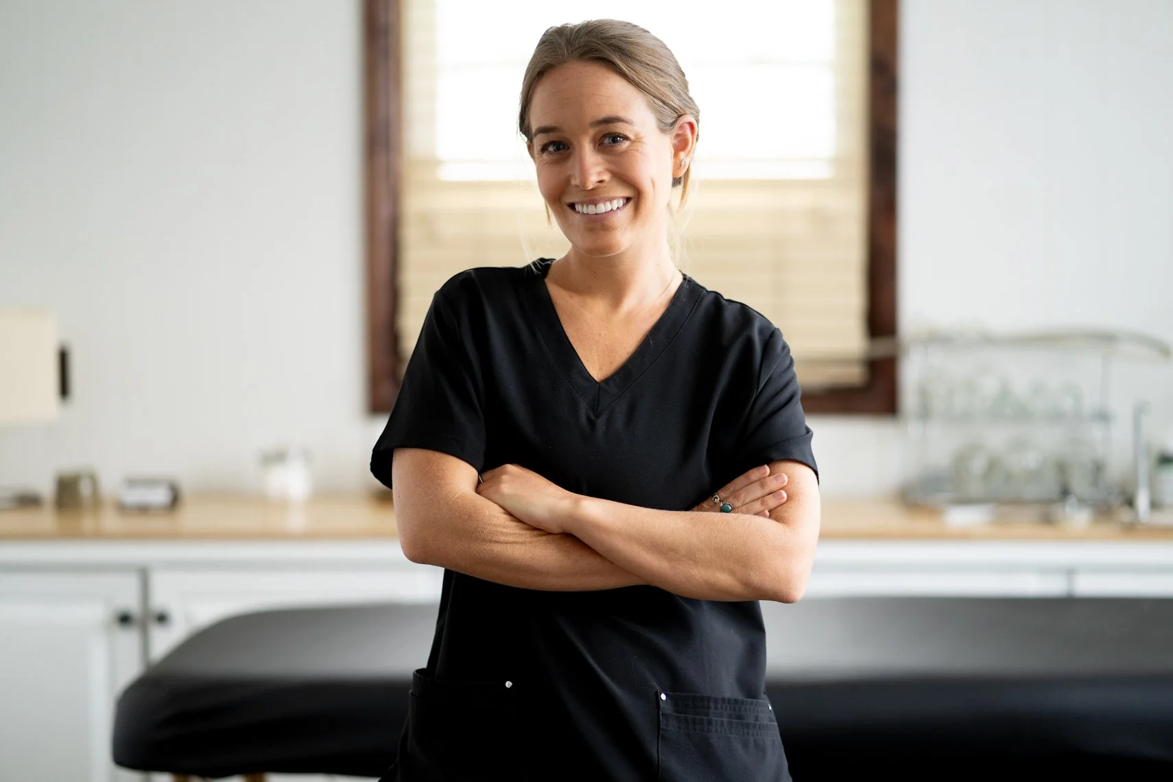 A smiling woman with light brown hair and wearing black medical scrubs standing with arms crossed in a clinical or massage practice setting with a window and countertop in the background.