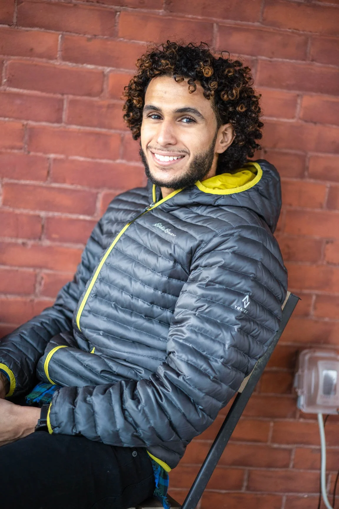 A smiling young man with curly hair and a beard, sitting on a chair in front of a red brick wall, wearing a gray and yellow puffy jacket.
