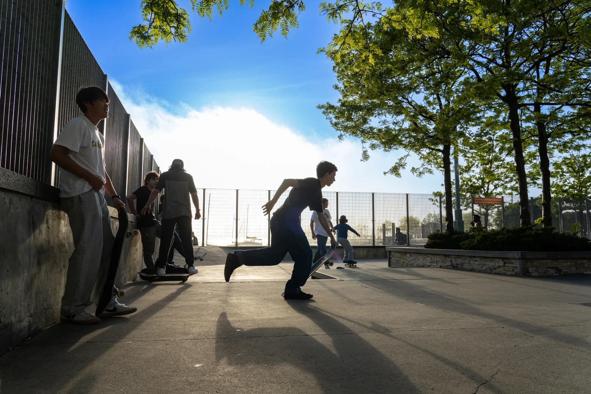 Group of young people at outdoor skatepark, skateboarding and socializing, with trees and a blue sky in the background.