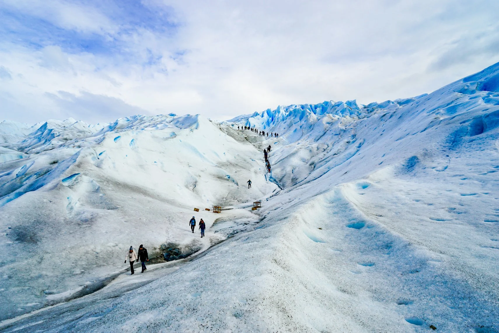 A group of people climbing on a glacier with snowy, icy peaks in the background under a partly cloudy sky.